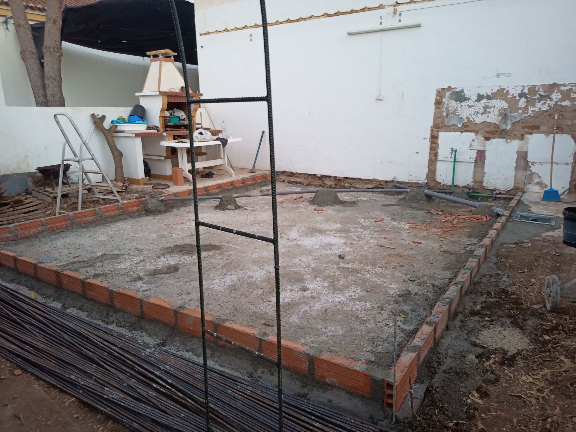 Canteiro de obras: Bordas de tijolos delimitam uma base de concreto. Vergalhões e uma escada estão presentes, com uma churrasqueira ao fundo.