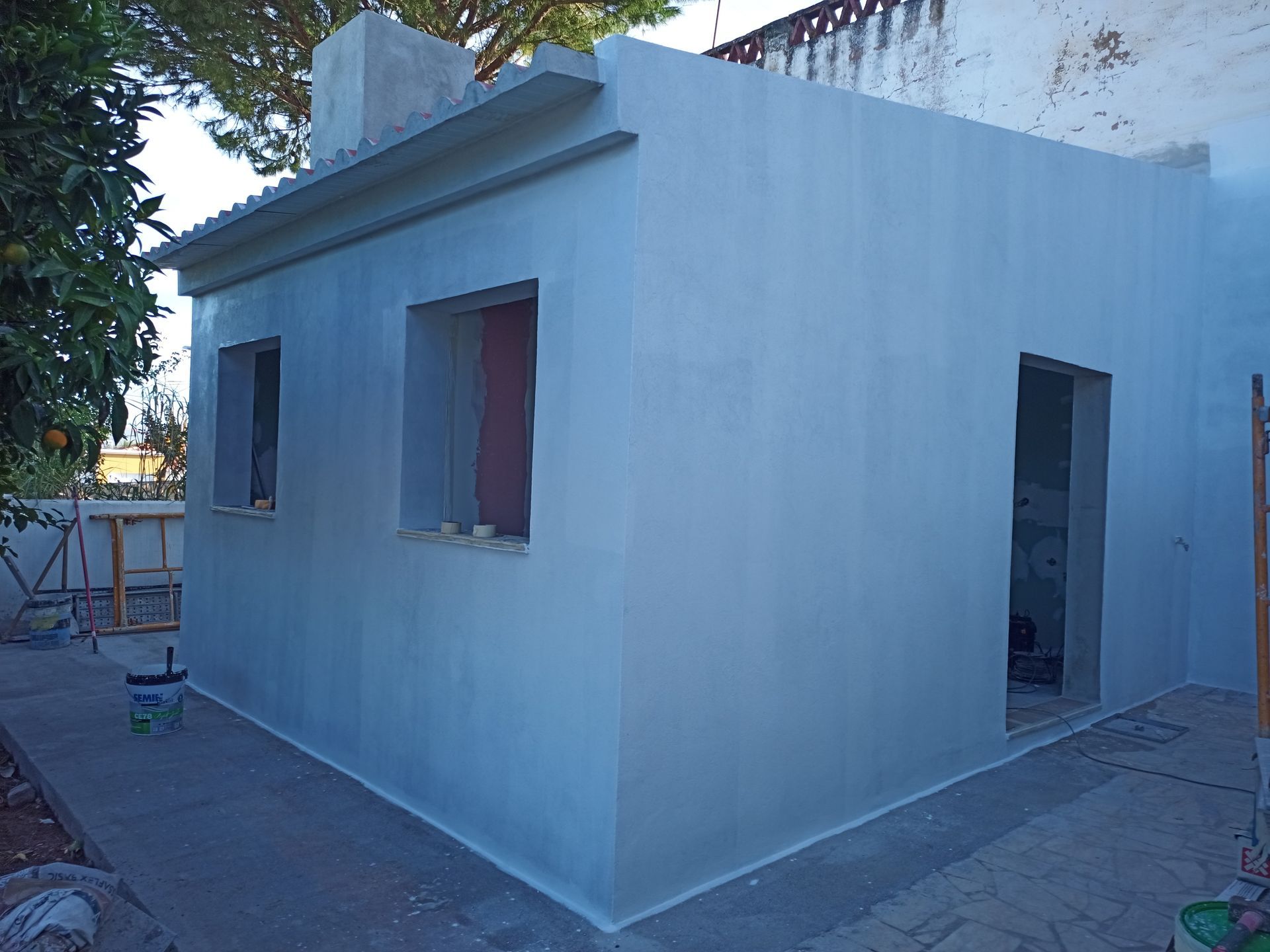 Exterior de um pequeno edifício em construção, paredes cinza-claro, porta aberta e caixilhos de janelas.