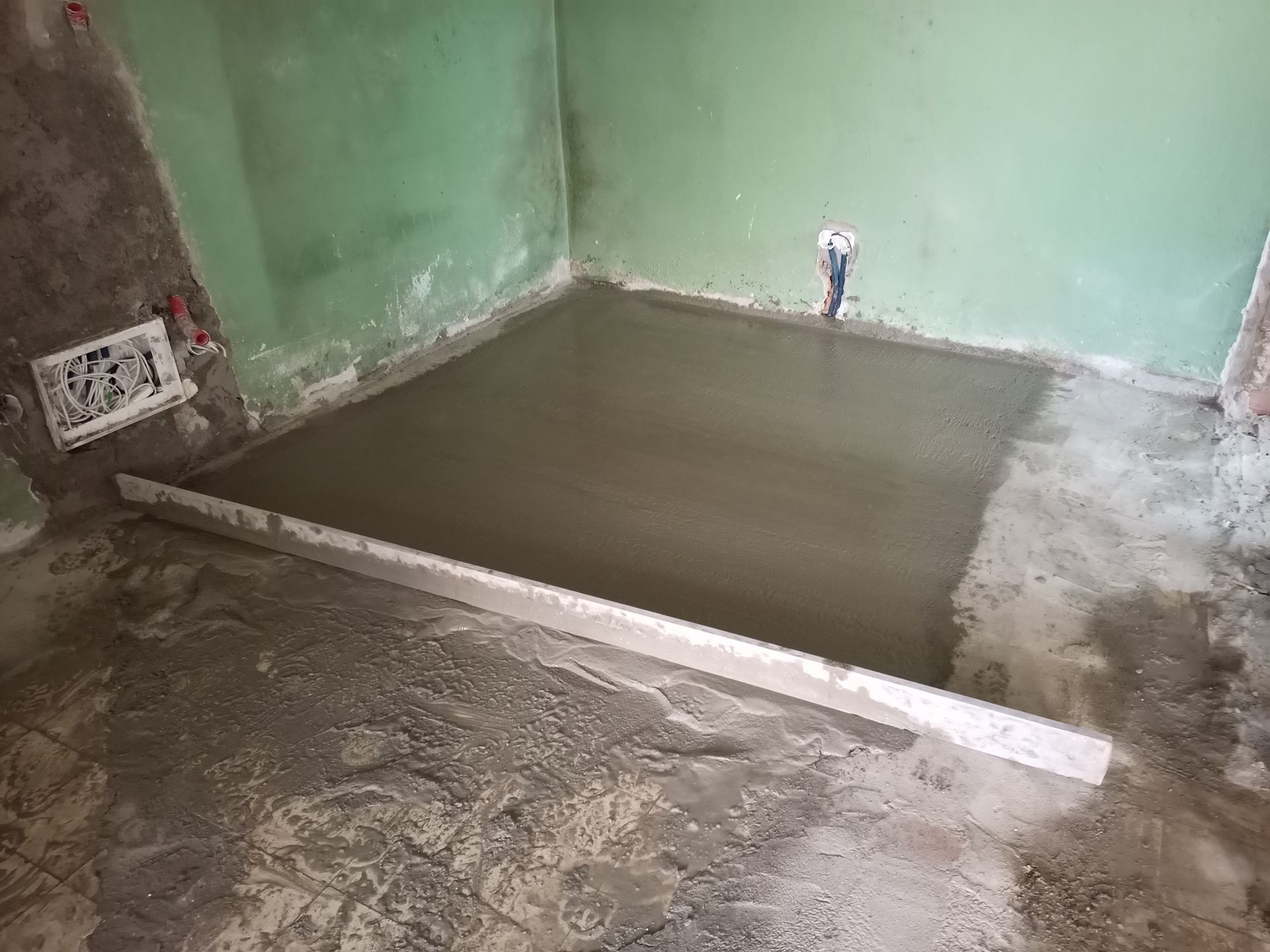 Base de chuveiro de concreto recém-moldada em um cômodo com paredes verdes e encanamento aparente.