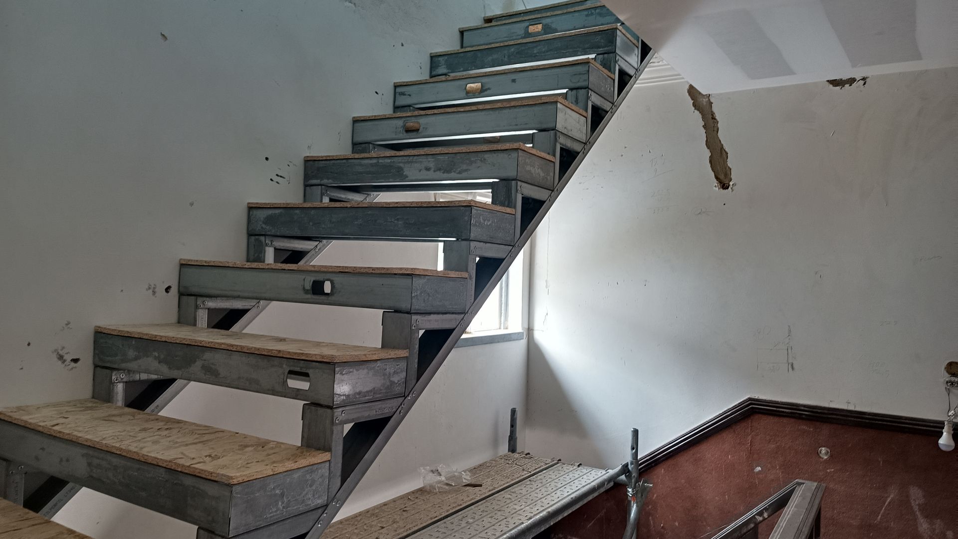 Escada metálica em um prédio com pintura descascando. A luz do dia entra por uma pequena janela lateral.