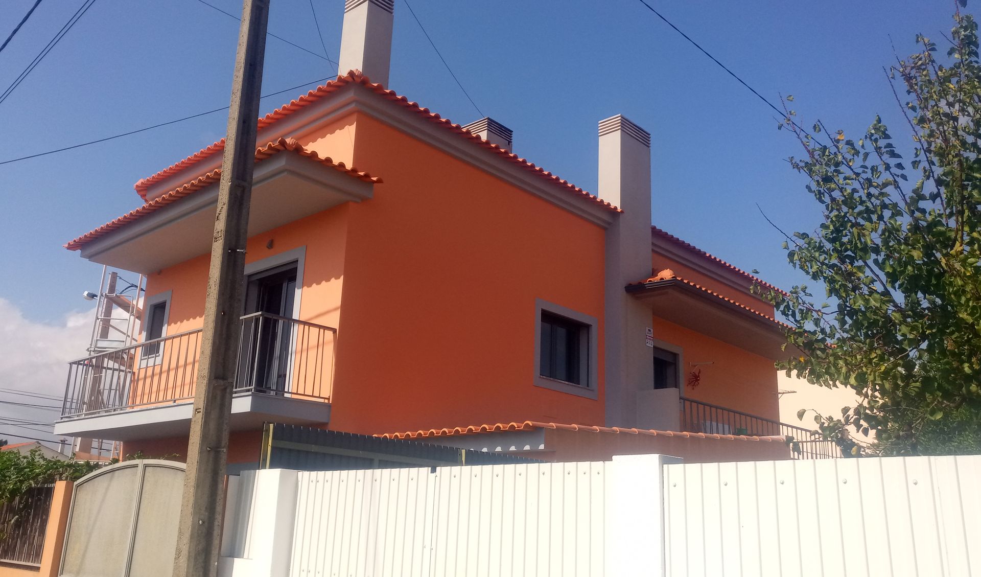 Casa laranja de dois andares com telhado vermelho, varanda e cerca branca sob um céu azul.