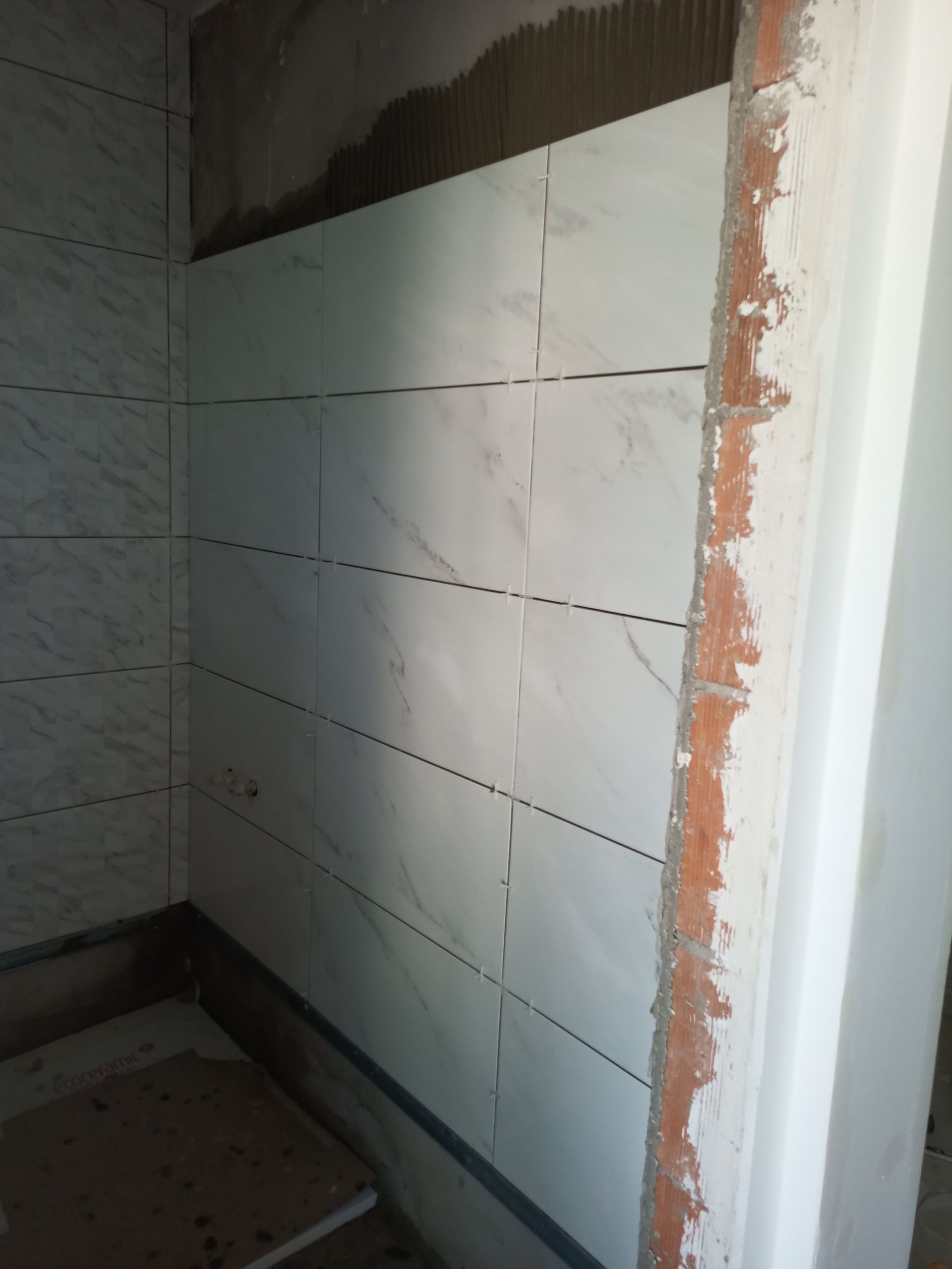 Azulejos retangulares brancos parcialmente instalados em uma parede de banheiro. Tijolos vermelhos e parede branca são visíveis.