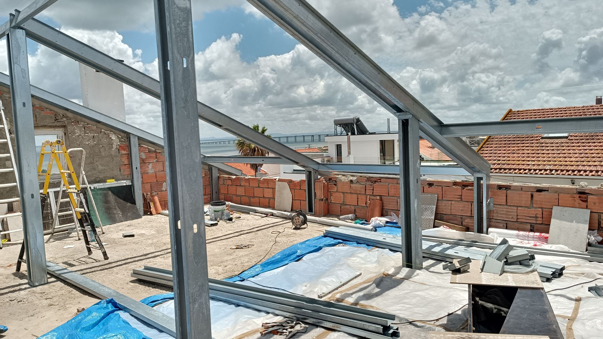 Estrutura de aço para um projeto de construção em cobertura com paredes de tijolo, lona azul e céu nublado.