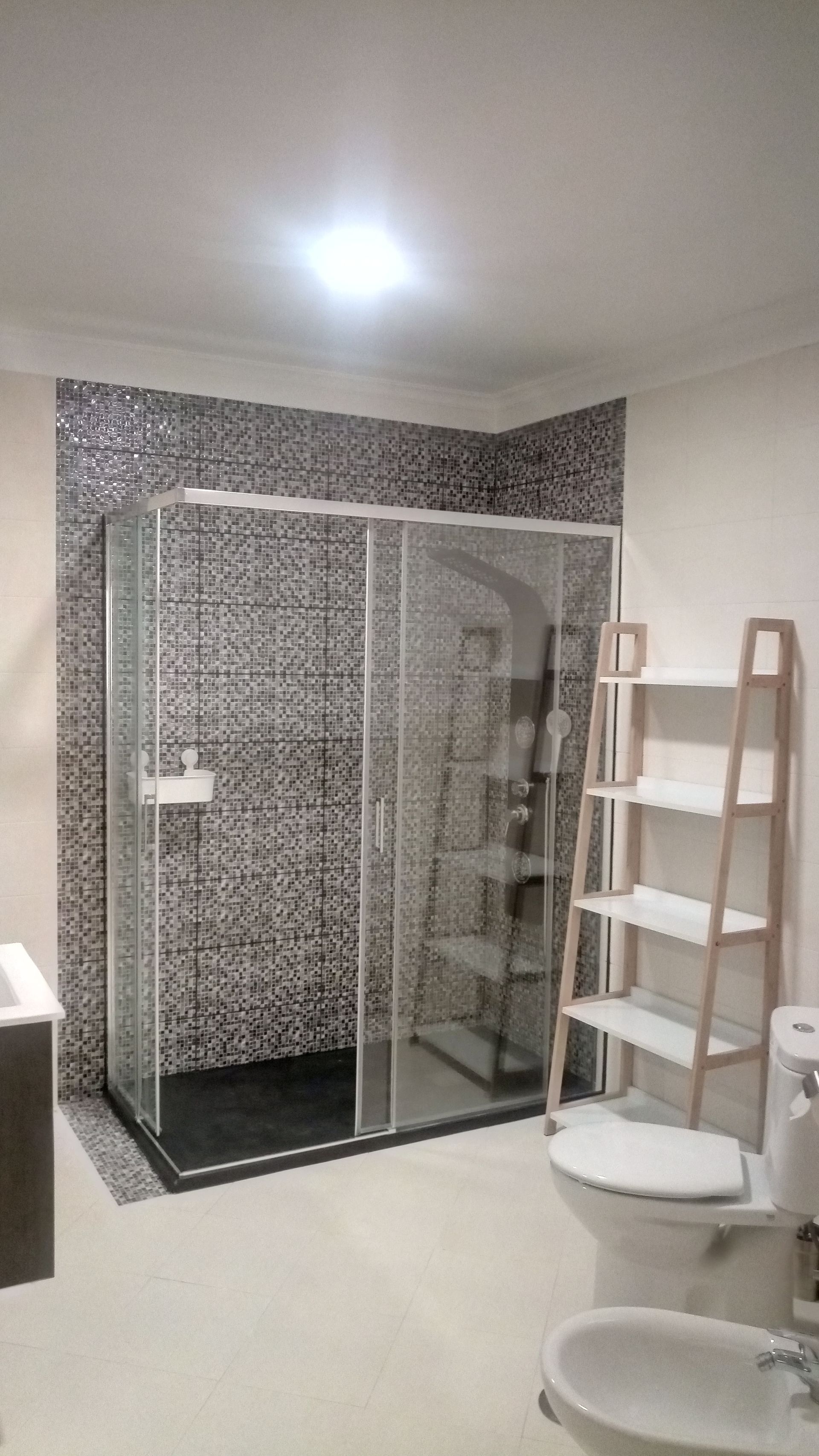 Banheiro moderno com box de vidro, parede revestida com azulejos em mosaico e prateleira de madeira.