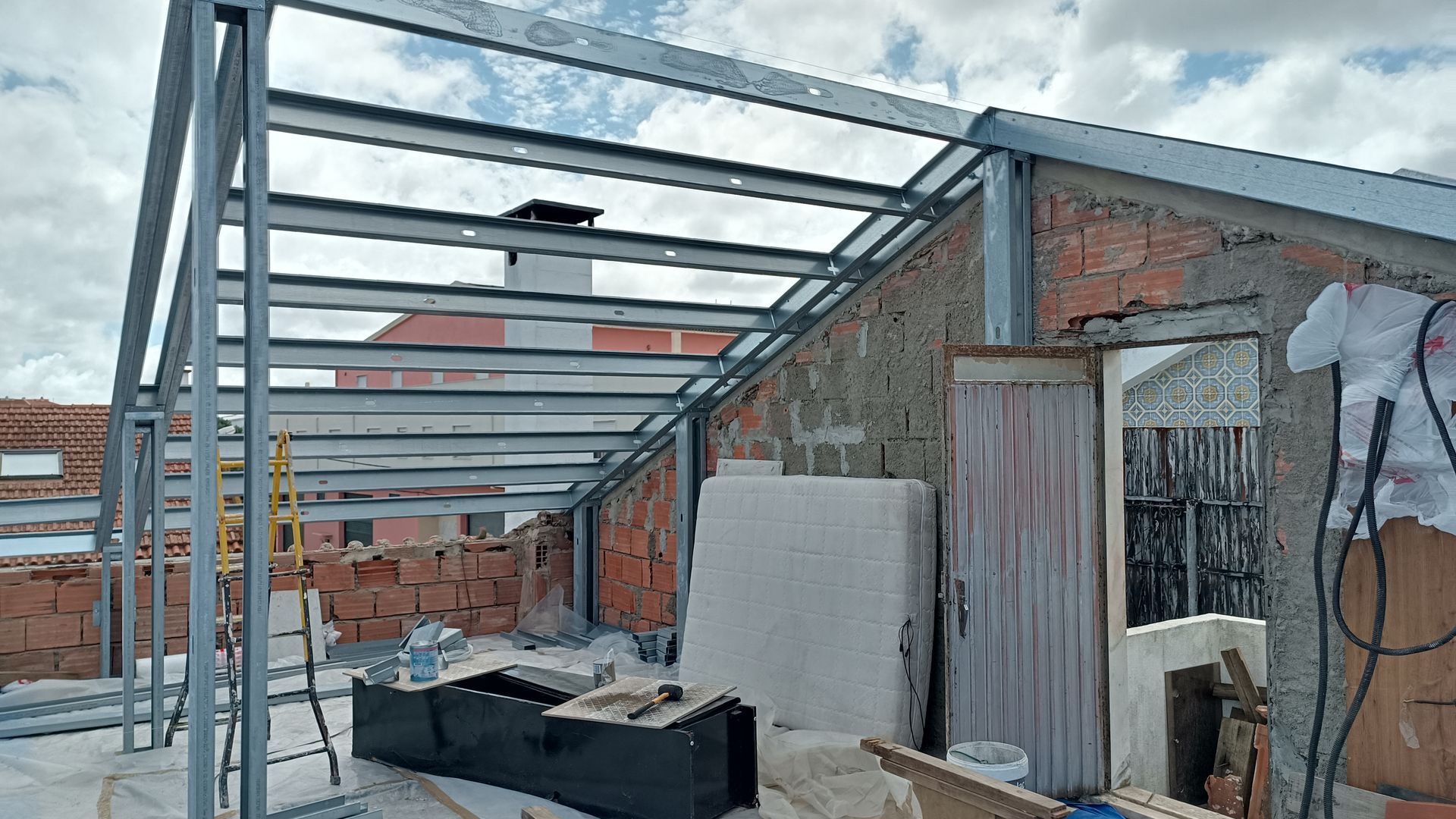 Estrutura metálica de um telhado em construção em um prédio de tijolos com céu nublado ao fundo.