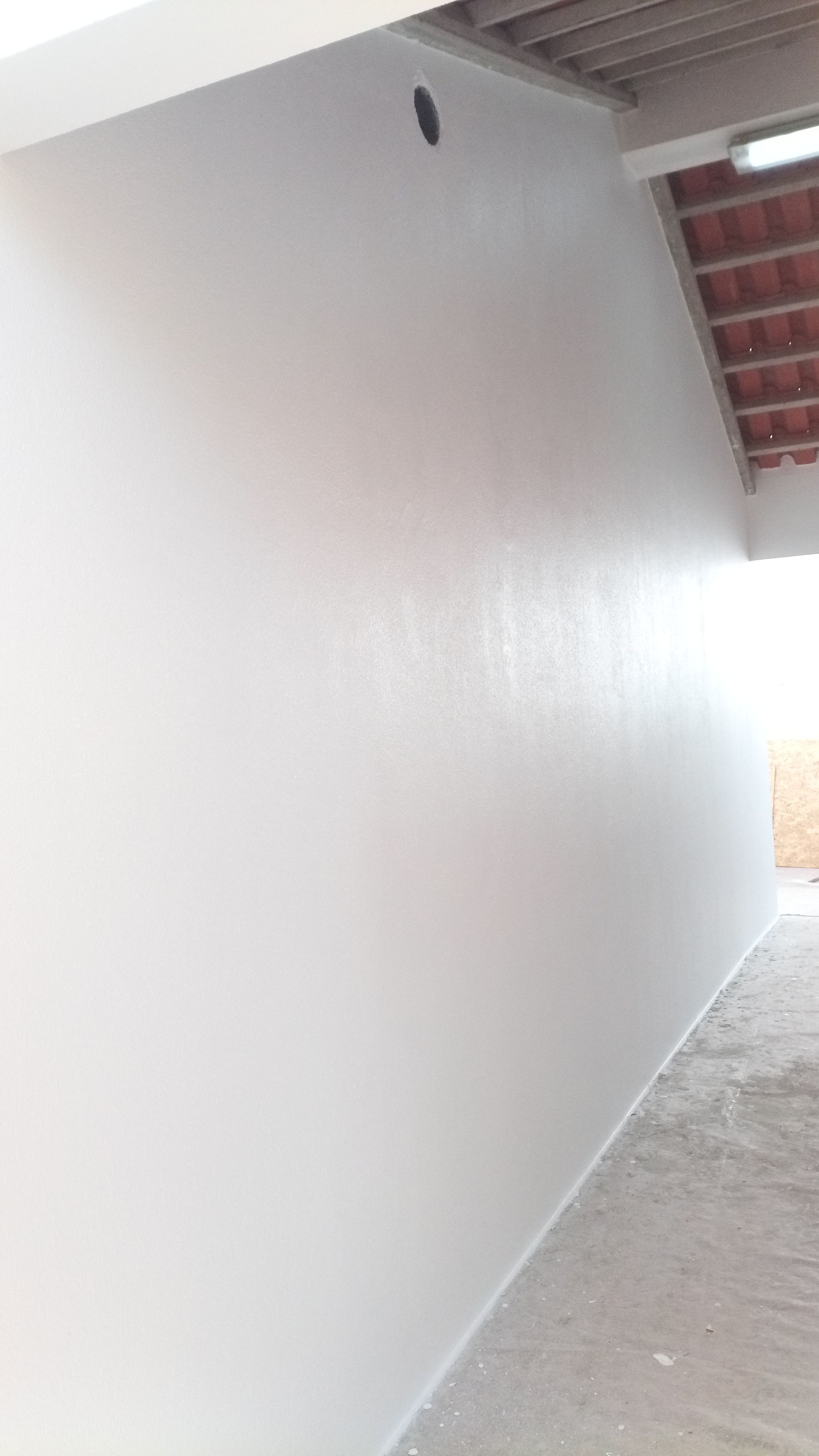 Parede pintada de branco sob um telhado com um pequeno buraco, piso de concreto.