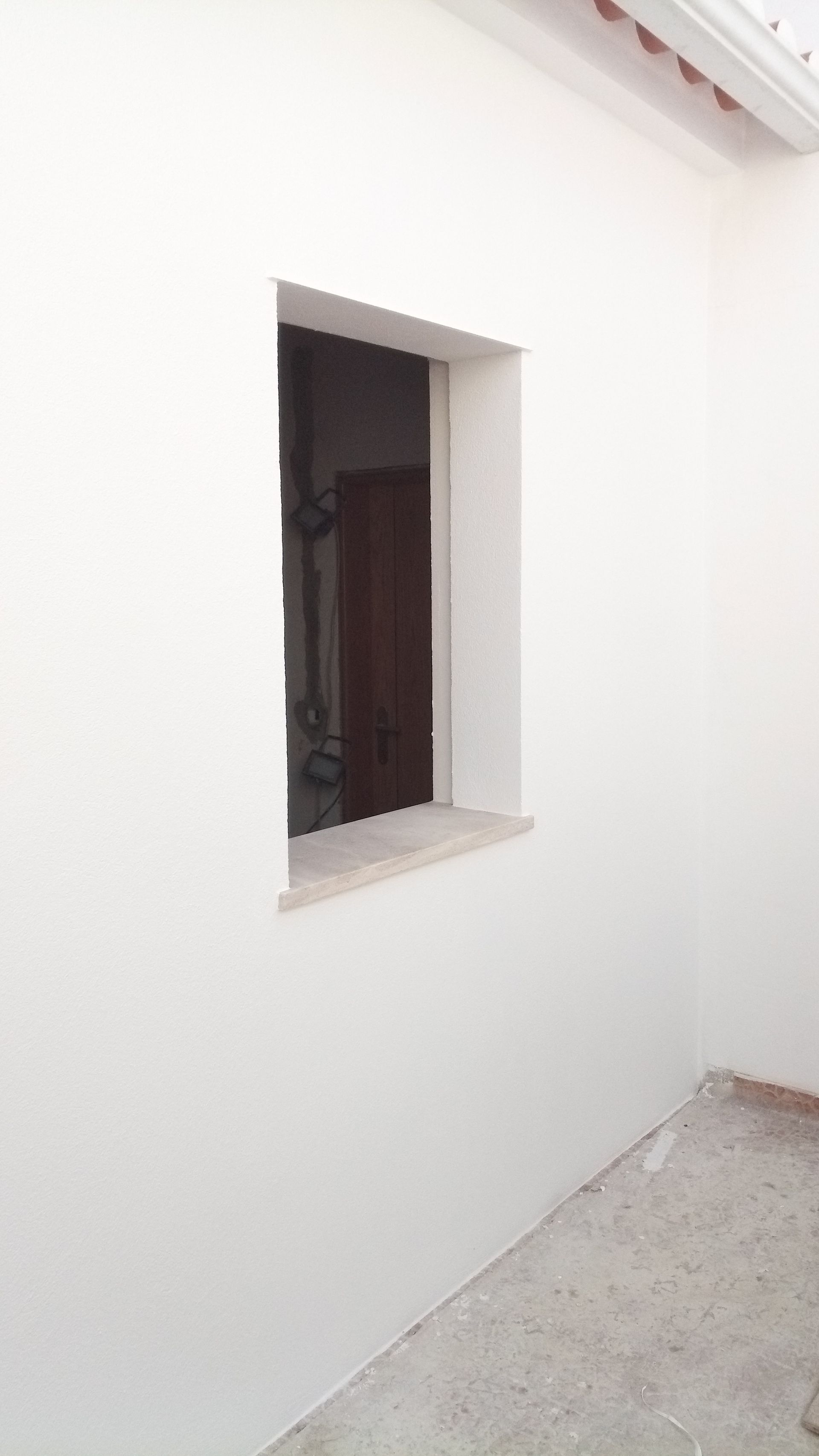Parede externa branca com uma pequena janela; uma porta é visível através da janela.
