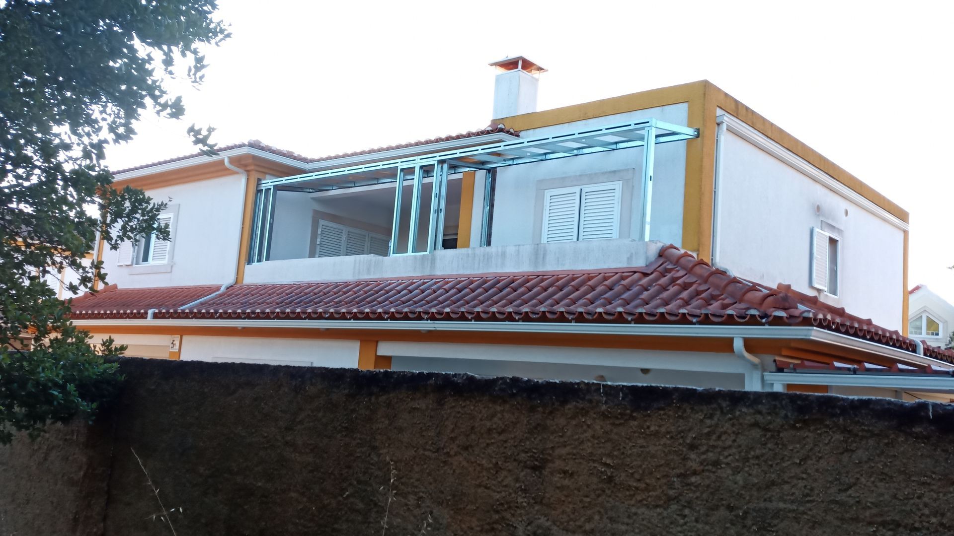 Casa de dois andares com telhado de telha vermelha, detalhes em amarelo e varanda envidraçada. Uma parede de concreto está em primeiro plano.