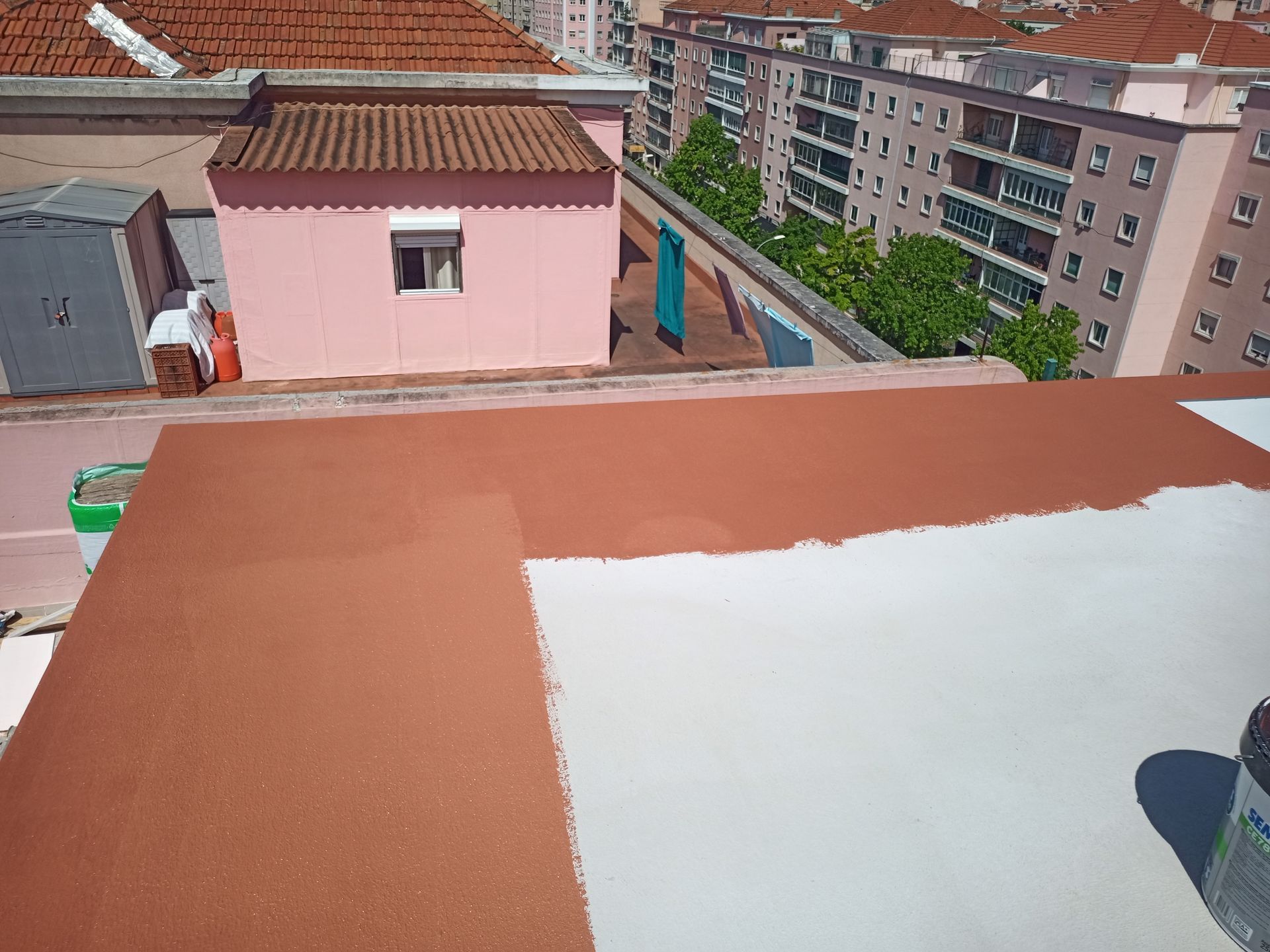 Telhado parcialmente pintado de marrom e branco, com prédios ao fundo. Uma lata de tinta é visível.
