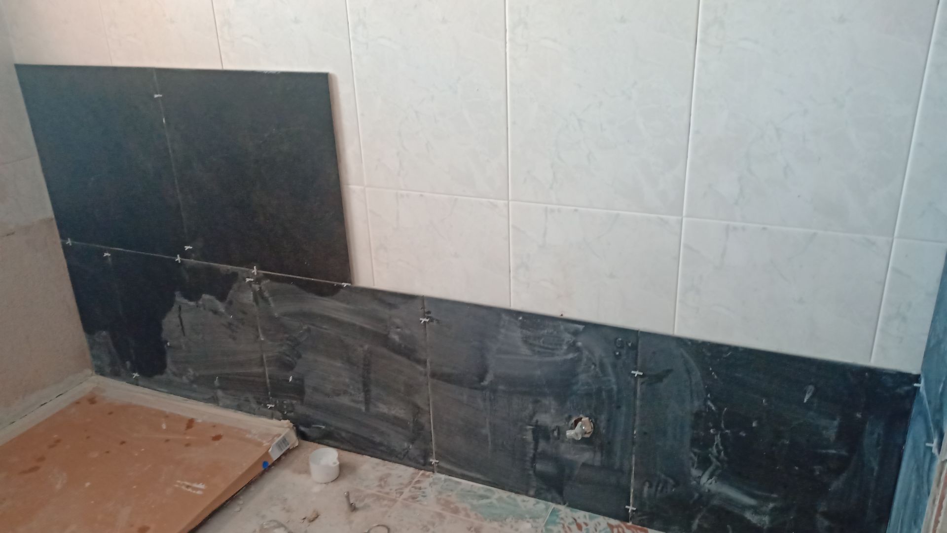Azulejos pretos sendo instalados em uma parede, com azulejos bege adjacentes. Argamassa visível nos azulejos instalados.