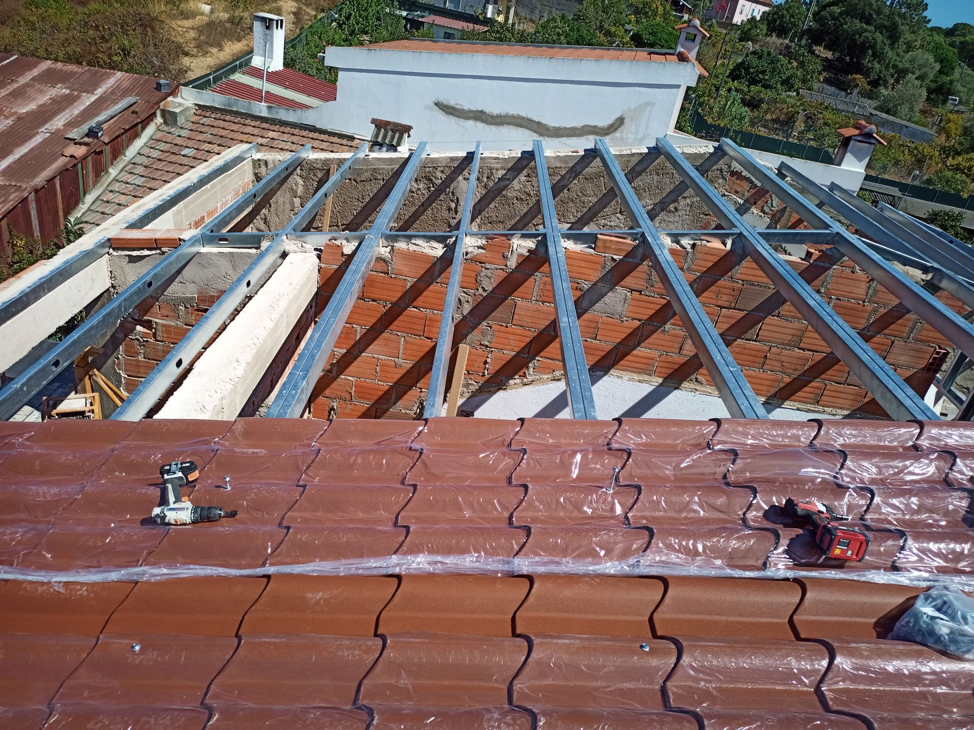 Telhado em construção: vigas metálicas sobre tijolo aparente, telhas em primeiro plano.