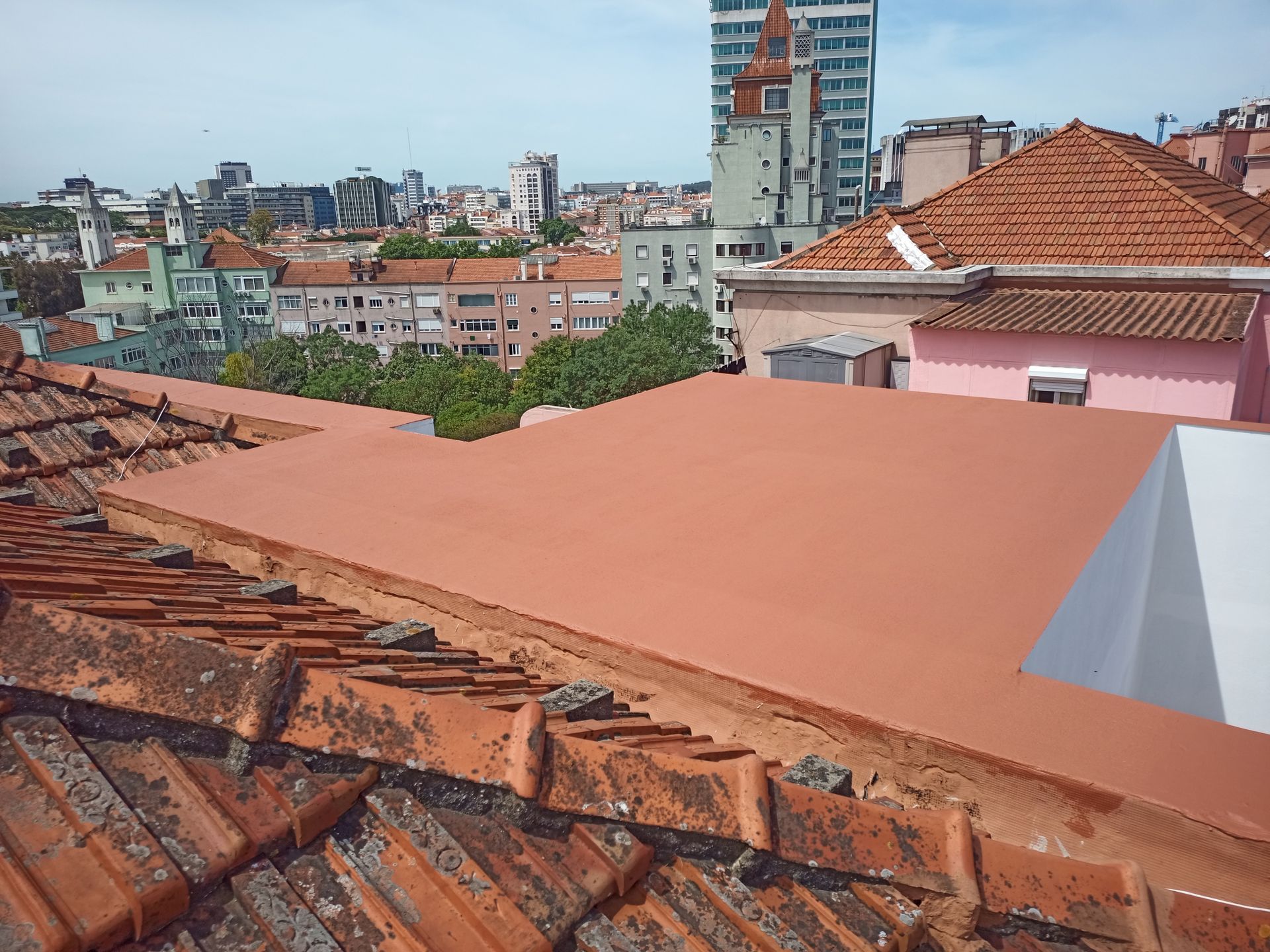 Vista dos telhados da cidade, com telhados de telha vermelha em primeiro plano, telhado plano laranja no meio e edifícios ao fundo sob um céu azul.