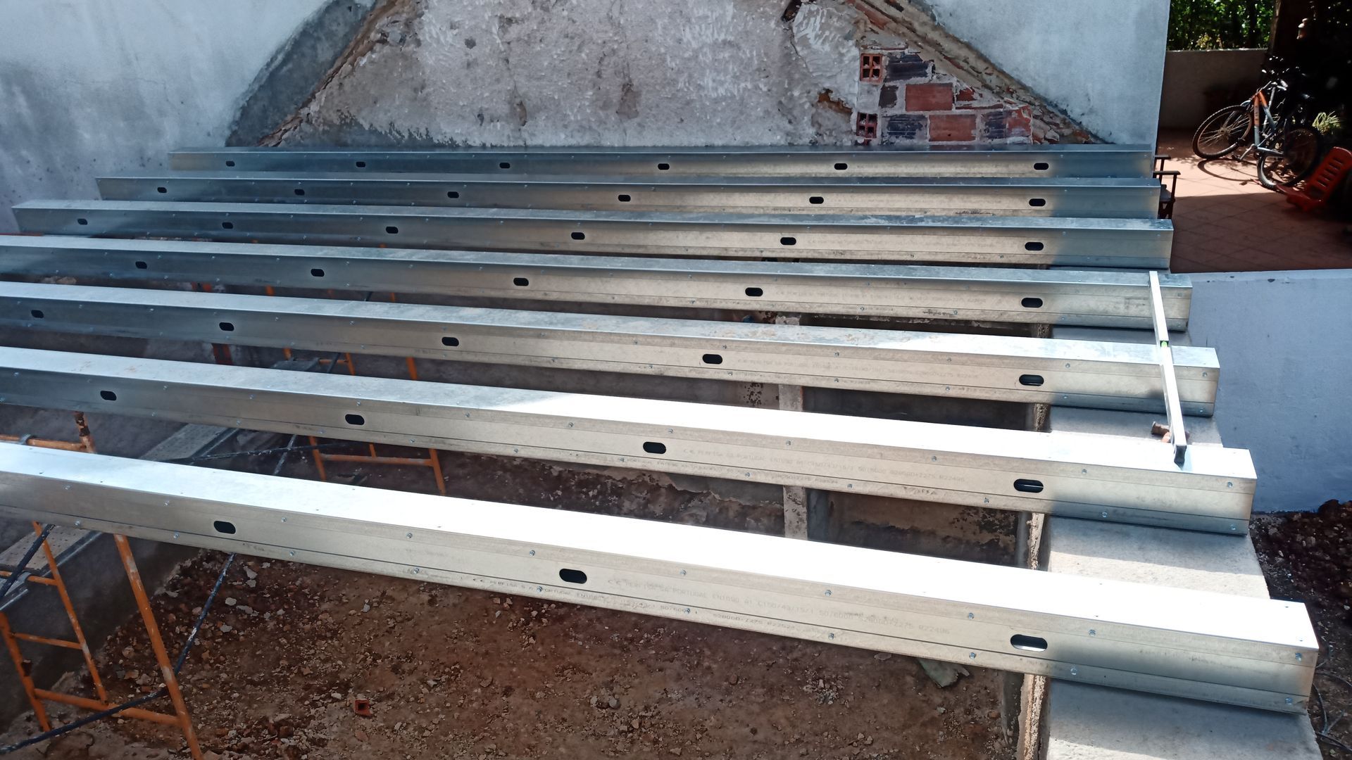 Vigas de metal empilhadas sobre uma superfície de concreto, perto de uma parede.