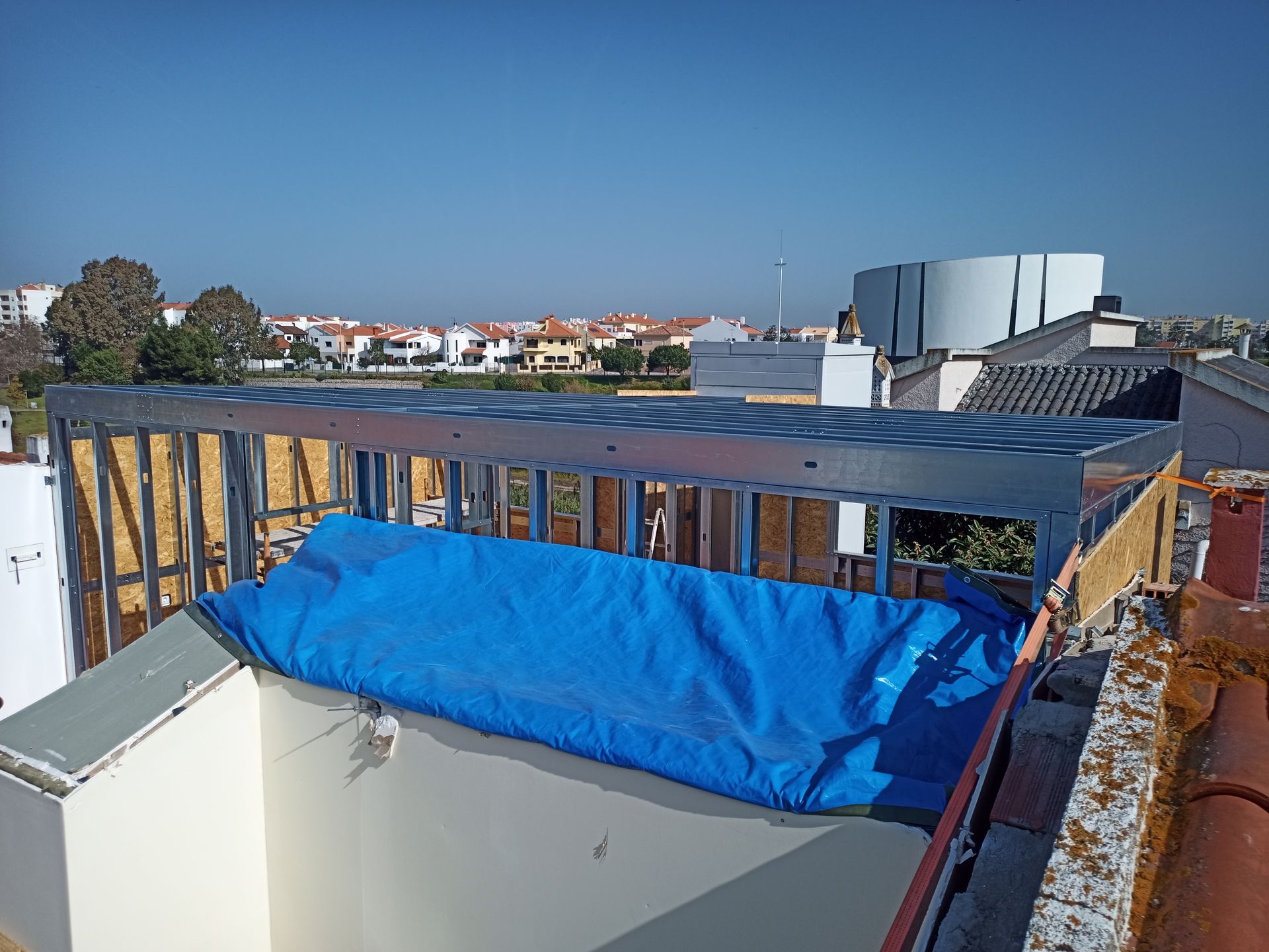 Estrutura metálica de um telhado em construção, coberta por uma lona azul, tendo como pano de fundo a paisagem urbana.