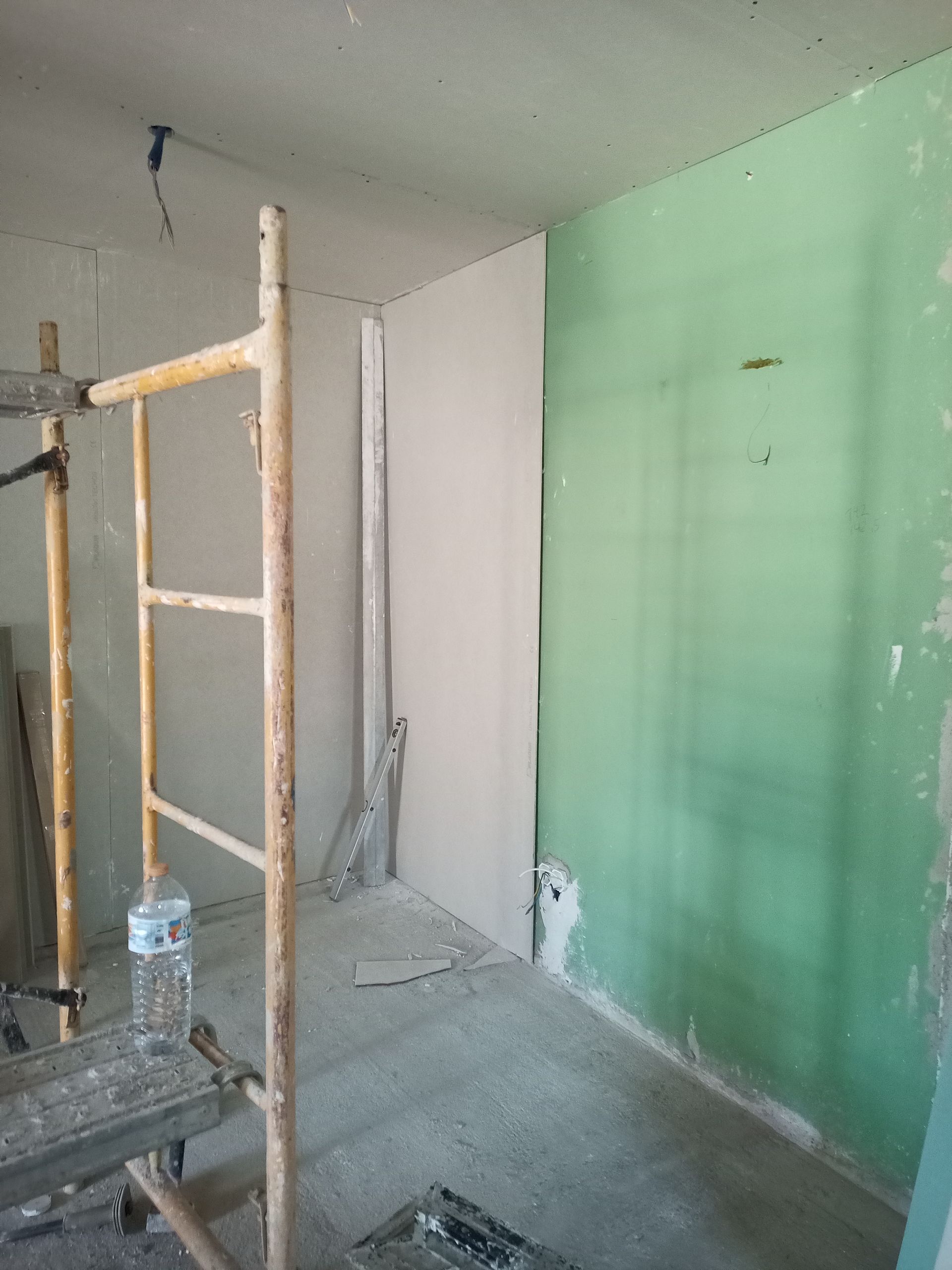Interior de uma obra com andaimes, paredes inacabadas em tons de cinza e verde, piso de concreto.