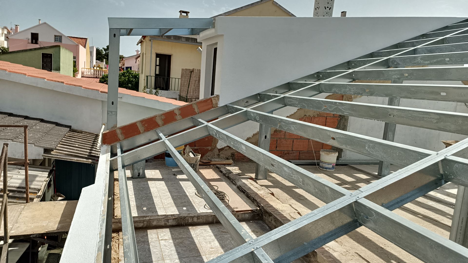Estrutura metálica do telhado em construção, com parte da cobertura em tijolo e edifícios circundantes.