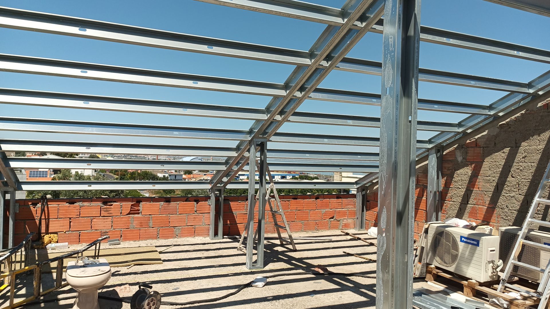 Estrutura metálica em construção em um telhado, sustentada por paredes de tijolos; céu azul visível.