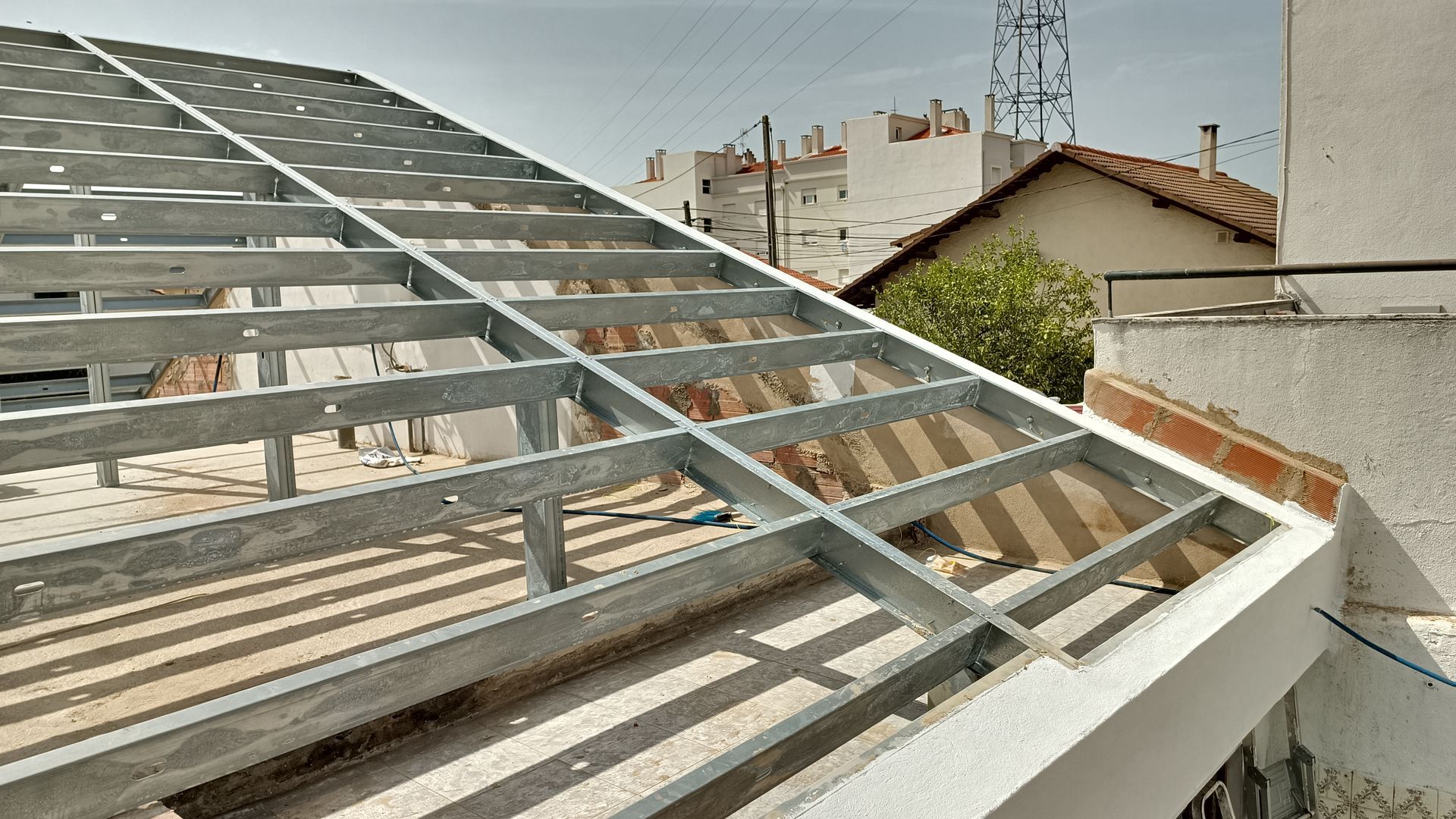 Estrutura metálica do telhado em construção. A luz do sol penetra pela estrutura; uma paisagem urbana se estende ao fundo.