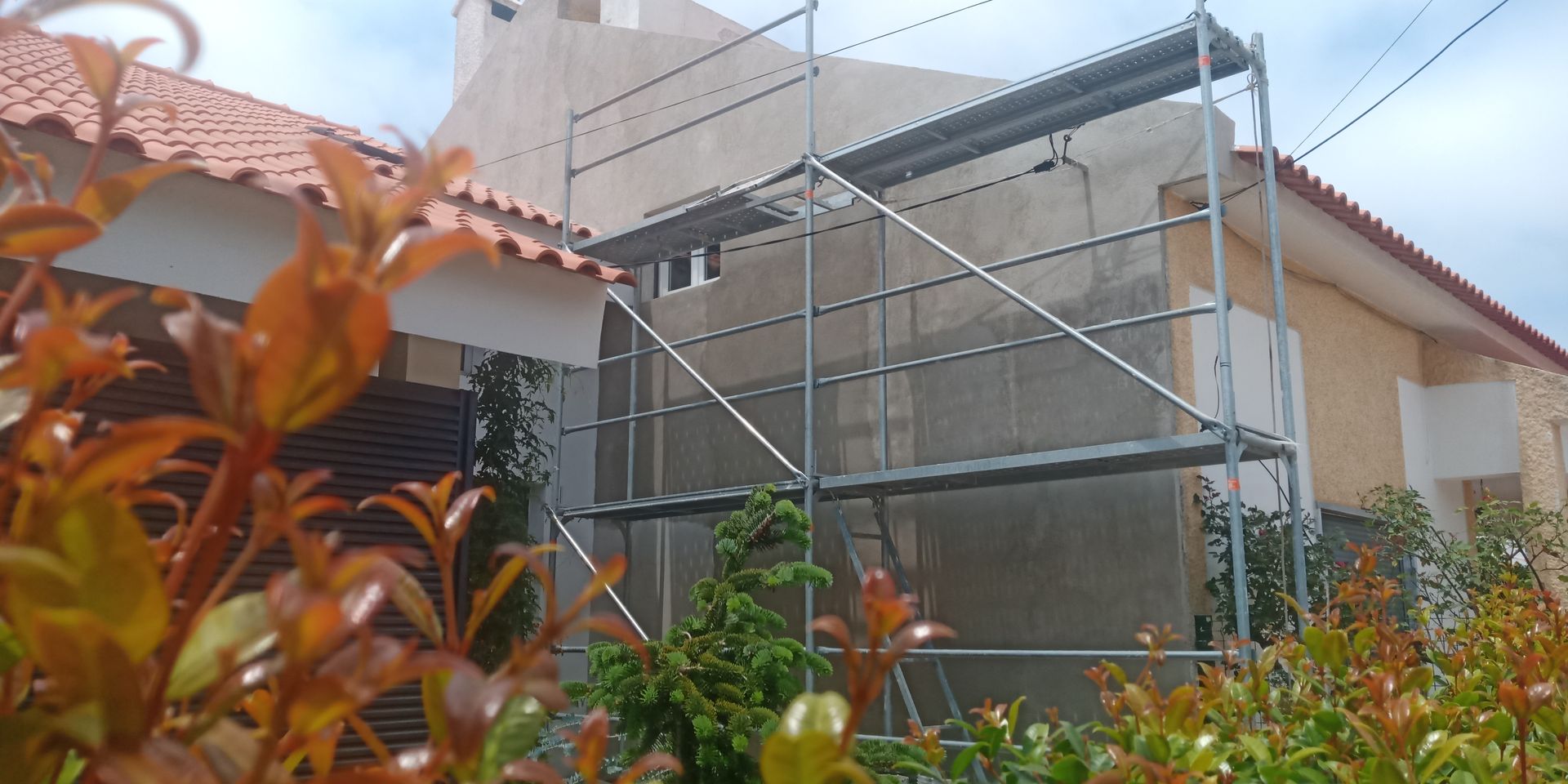 Um andaime se apoia na fachada bege de um prédio, cercado por folhagem verde e laranja sob um céu nublado.