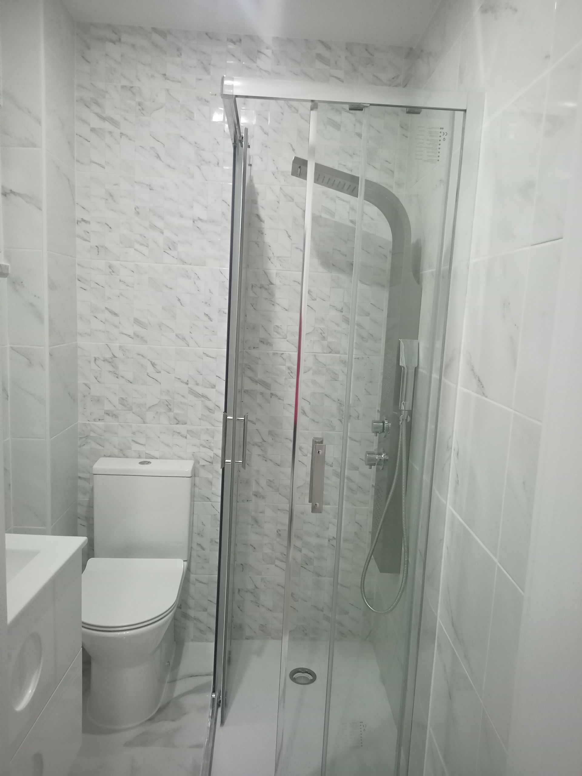 Banheiro com azulejos brancos, vaso sanitário e box de vidro para o chuveiro.