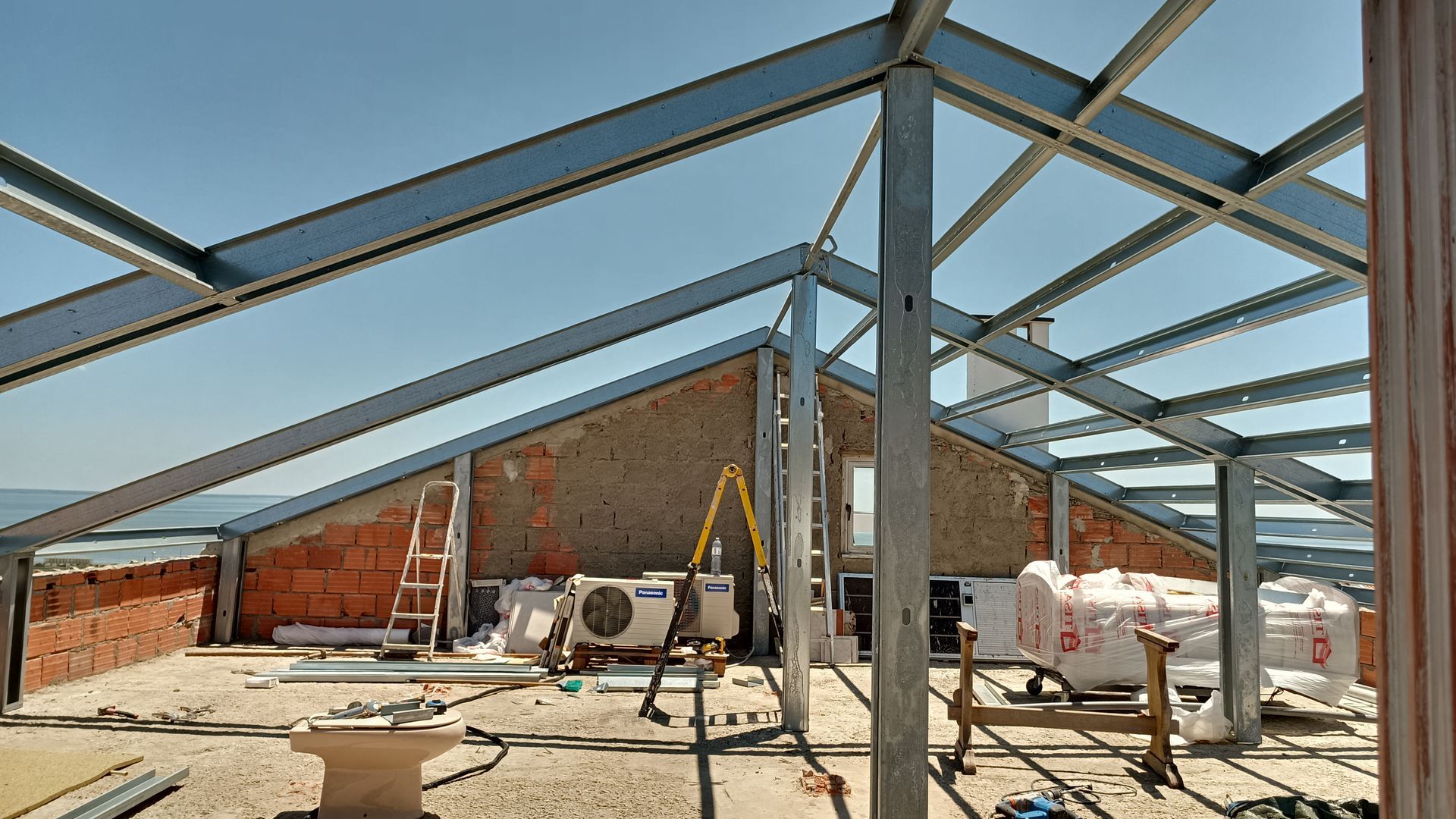 Estrutura metálica de um telhado em construção contra um céu azul, com paredes de tijolo e equipamentos em um telhado plano.