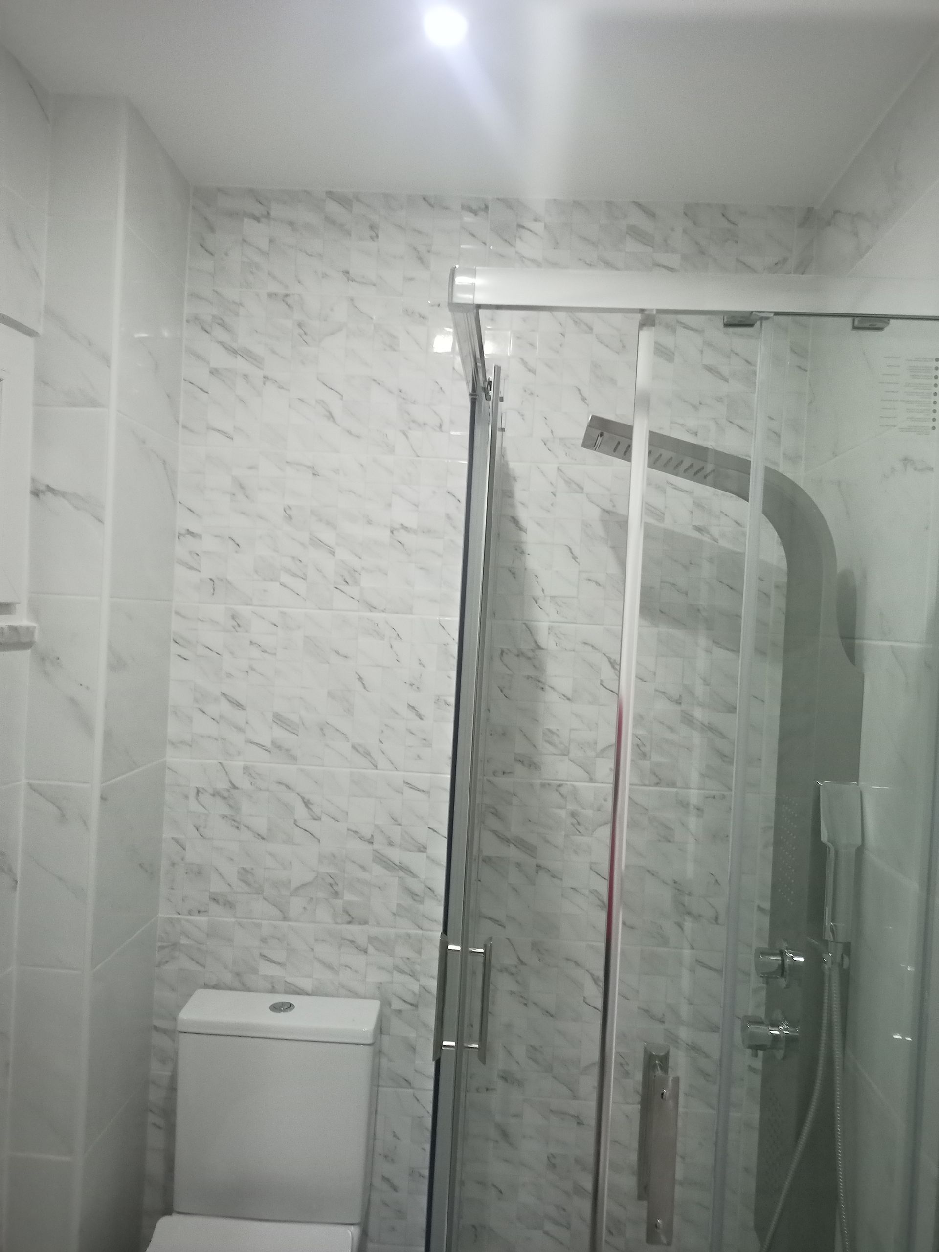 Interior do banheiro com azulejos com estampa de mármore, box de vidro, vaso sanitário e luminária de teto.