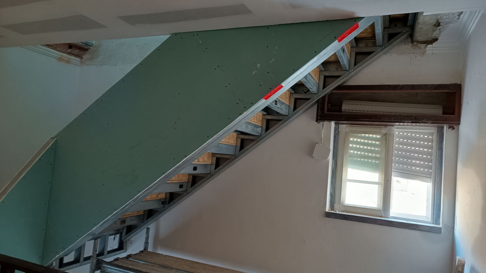 Escada em construção com estrutura metálica aparente, drywall e pequena janela. O drywall verde contrasta com o metal e as paredes brancas.