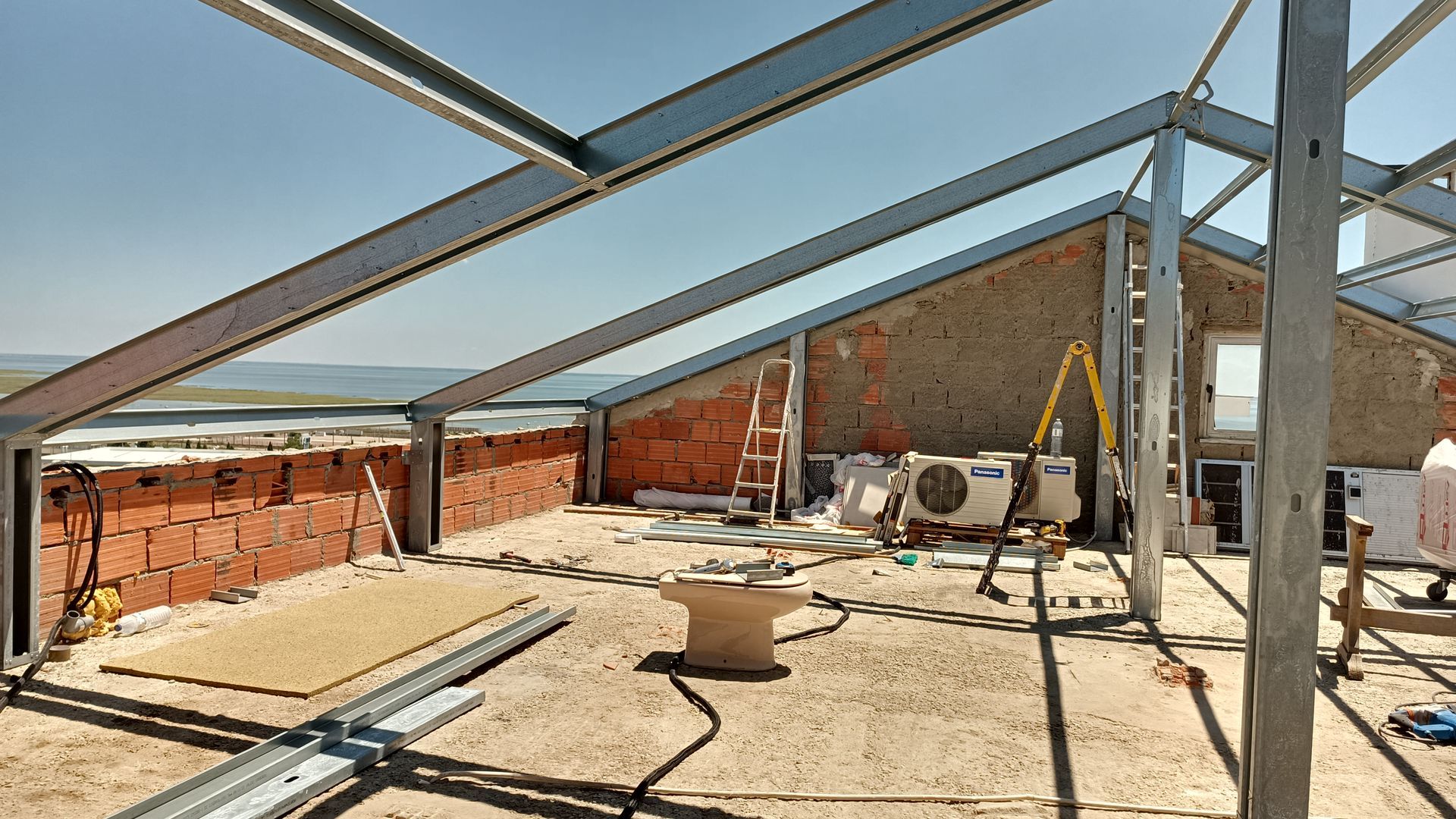 Canteiro de obras: estrutura metálica, paredes de tijolo, céu limpo, vista para o litoral. Interior inacabado.