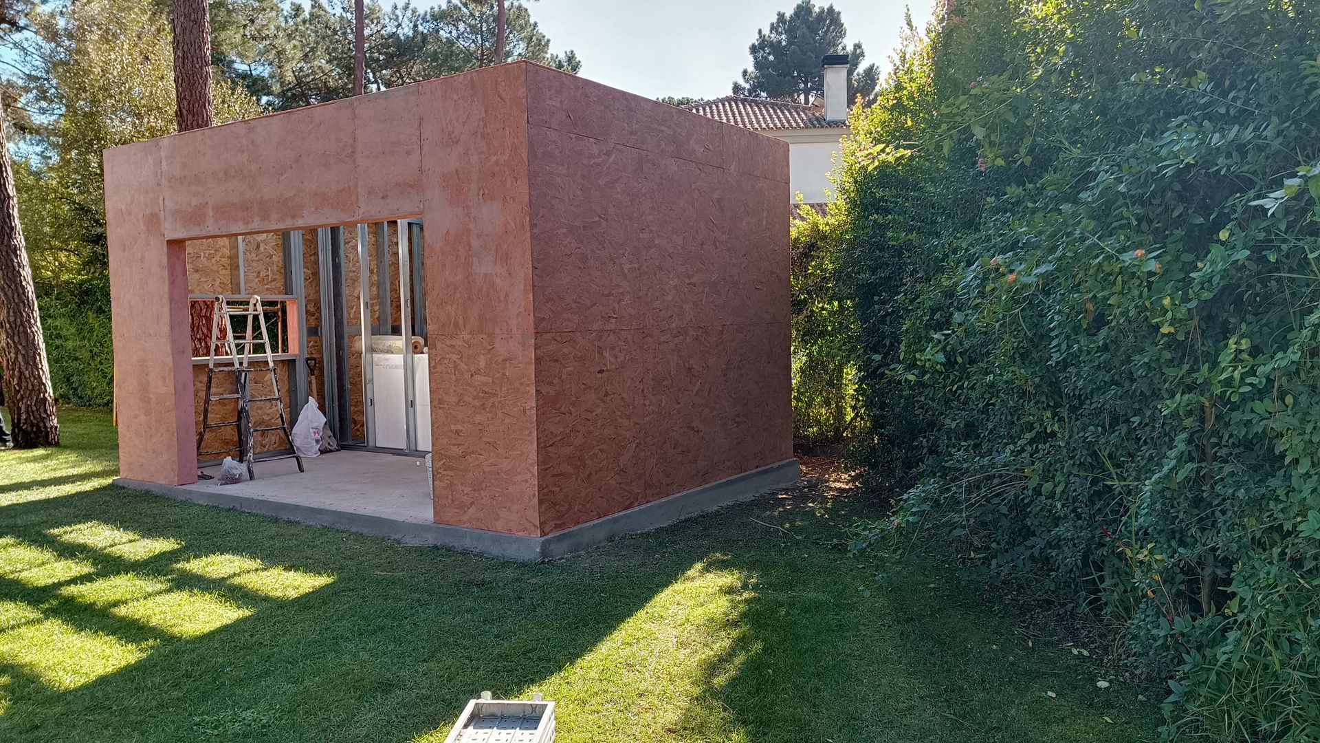 Um barracão parcialmente construído, com exterior marrom-avermelhado, frente aberta e cercado por grama verde e arbustos.