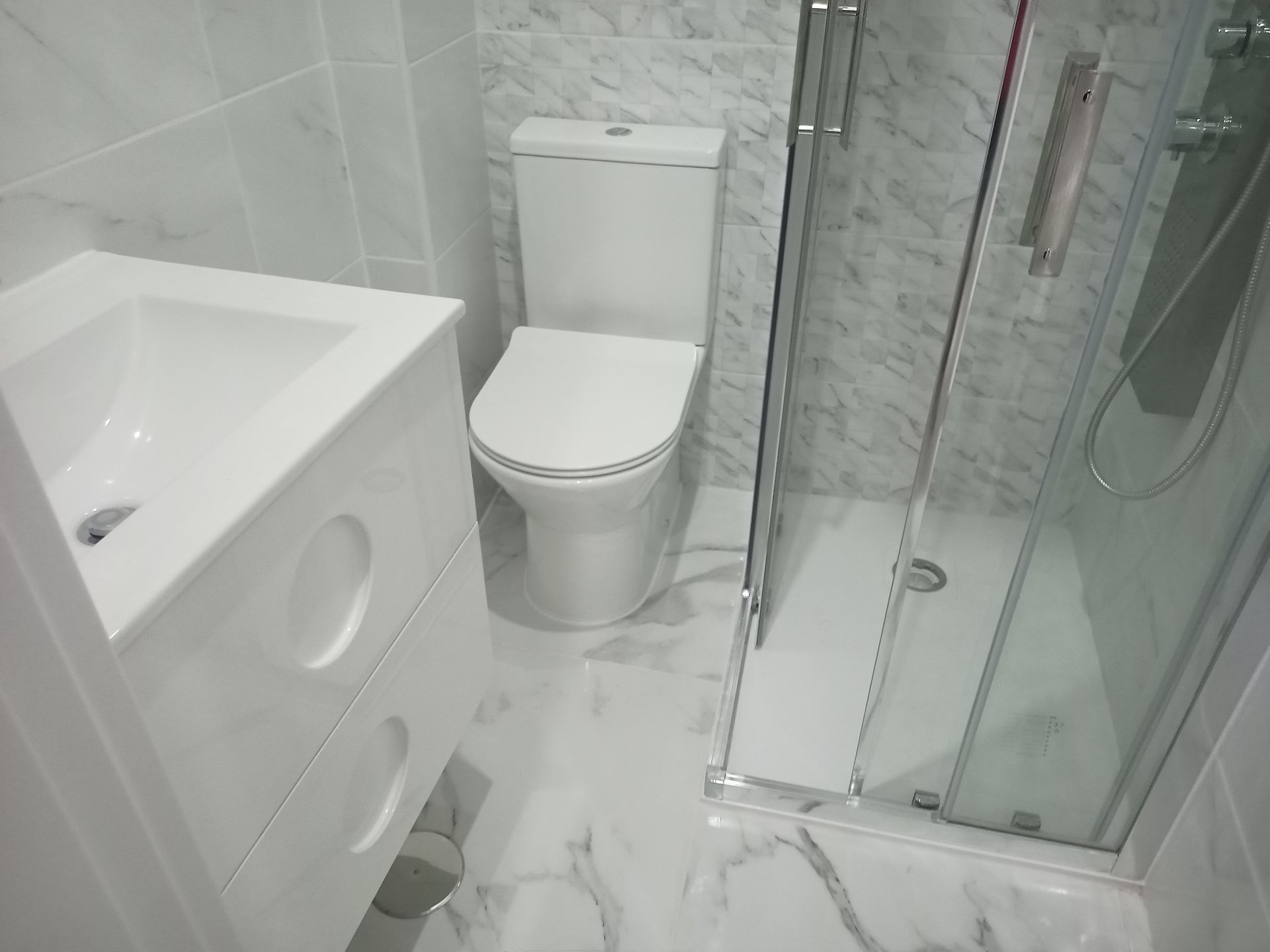 Pequeno banheiro branco com pia, vaso sanitário e chuveiro.