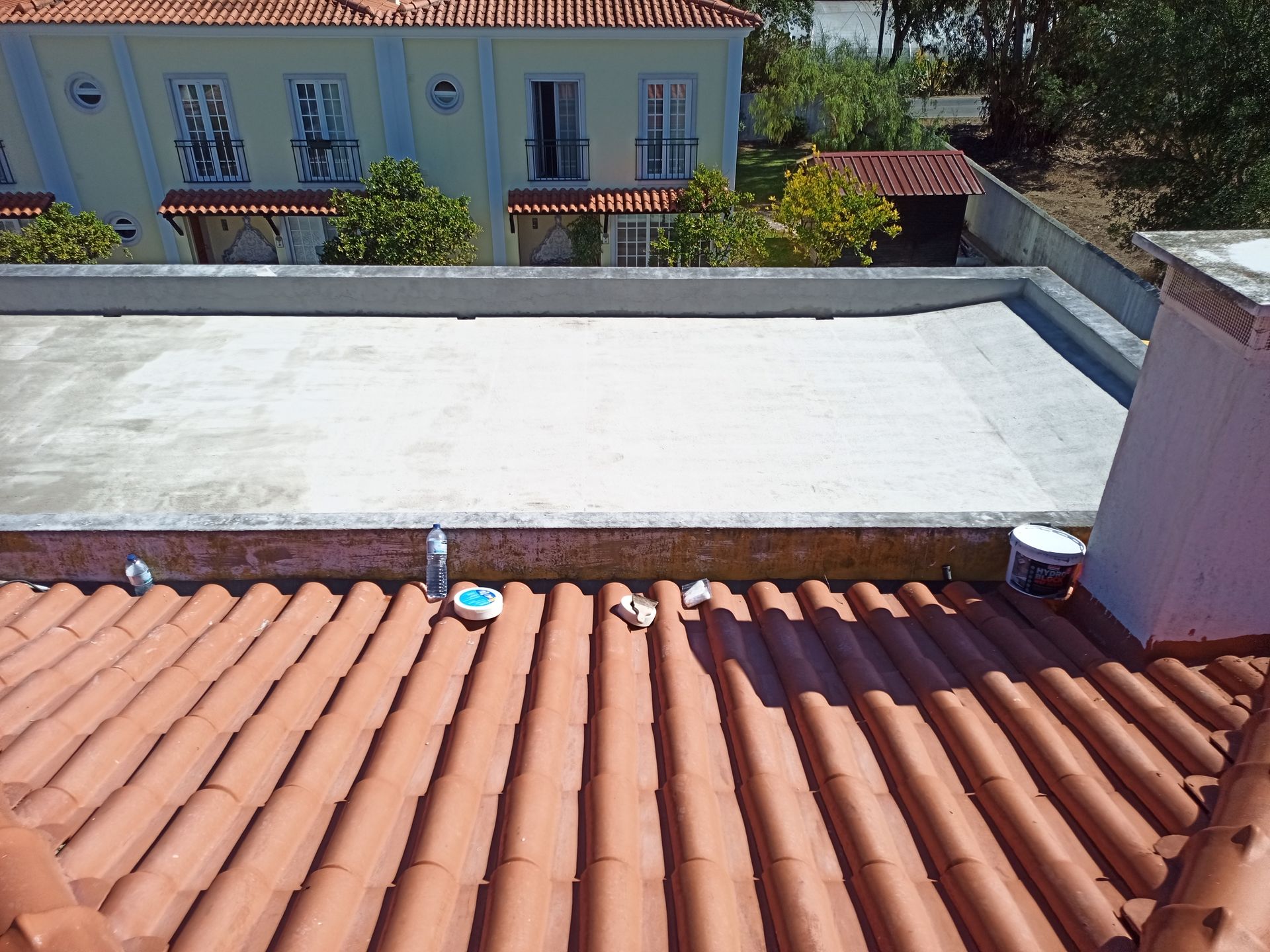 Telhado de telha vermelha em primeiro plano, com superfície de concreto e prédio ao fundo.