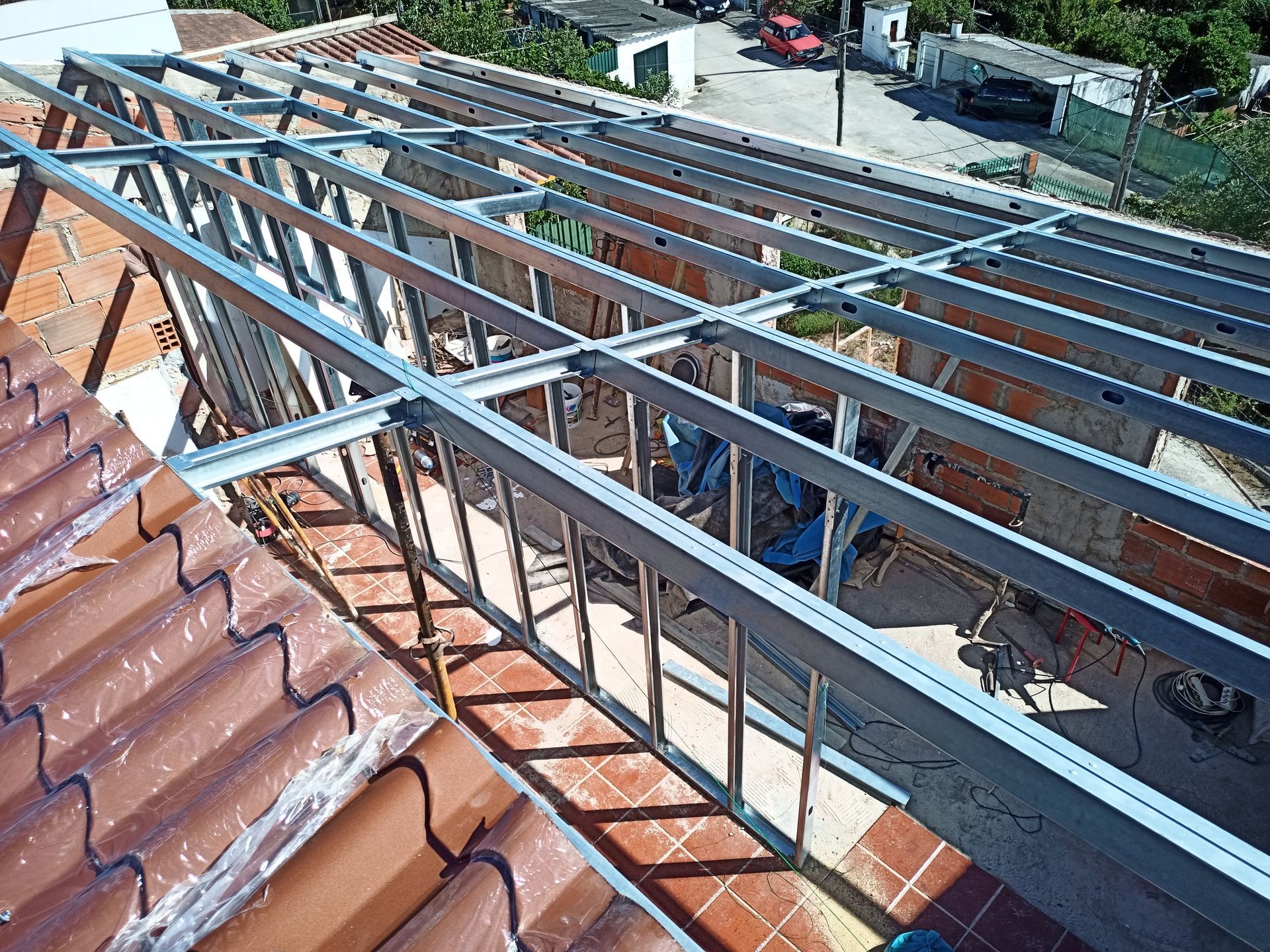 Estrutura metálica sobre um telhado de telhas vermelhas, com a luz do sol projetando sombras e uma área residencial ao fundo.
