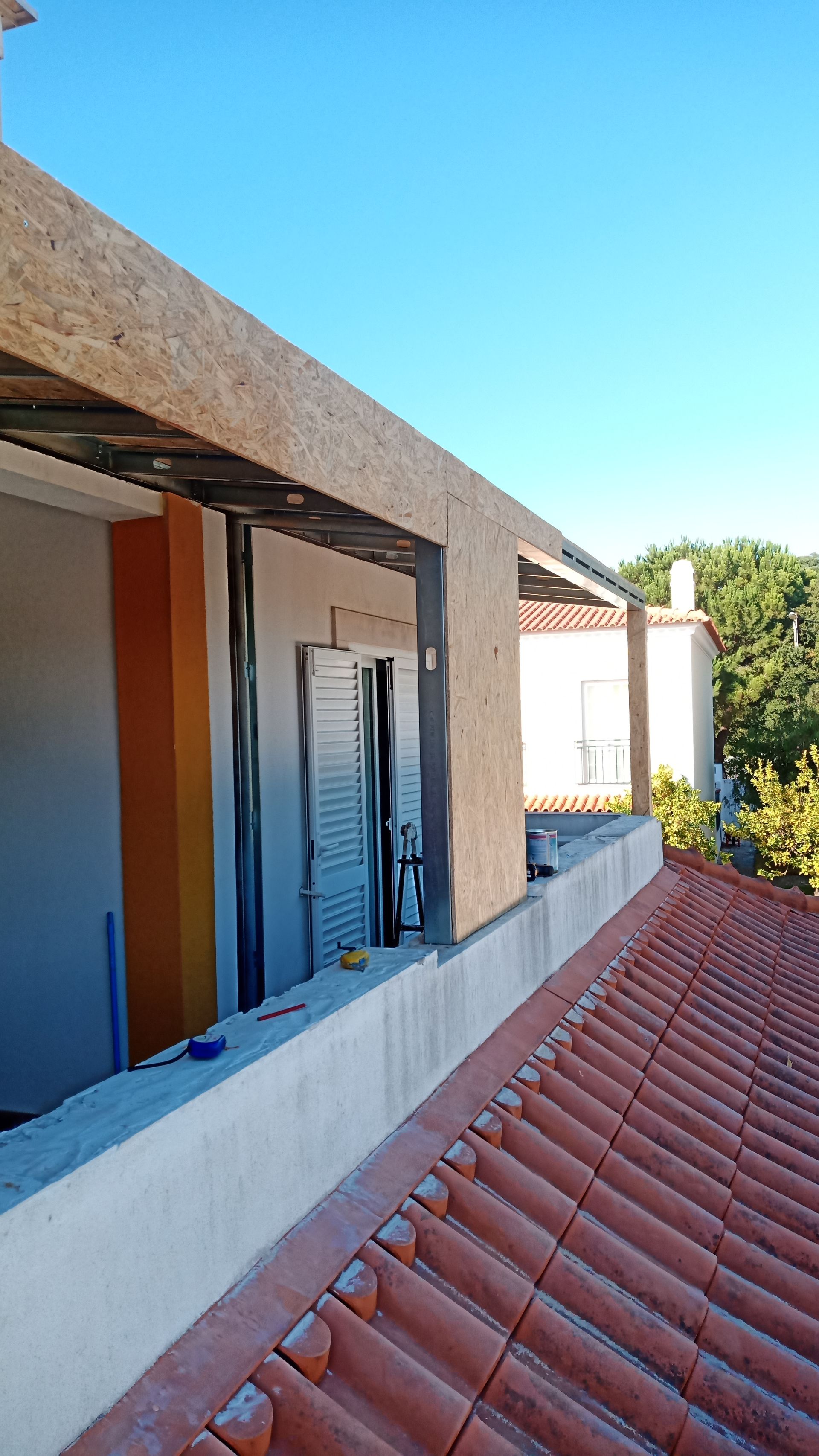 Vista do telhado da construção; estrutura de madeira acima da janela, paredes laranja e brancas, céu azul, telhado de telhas vermelhas.