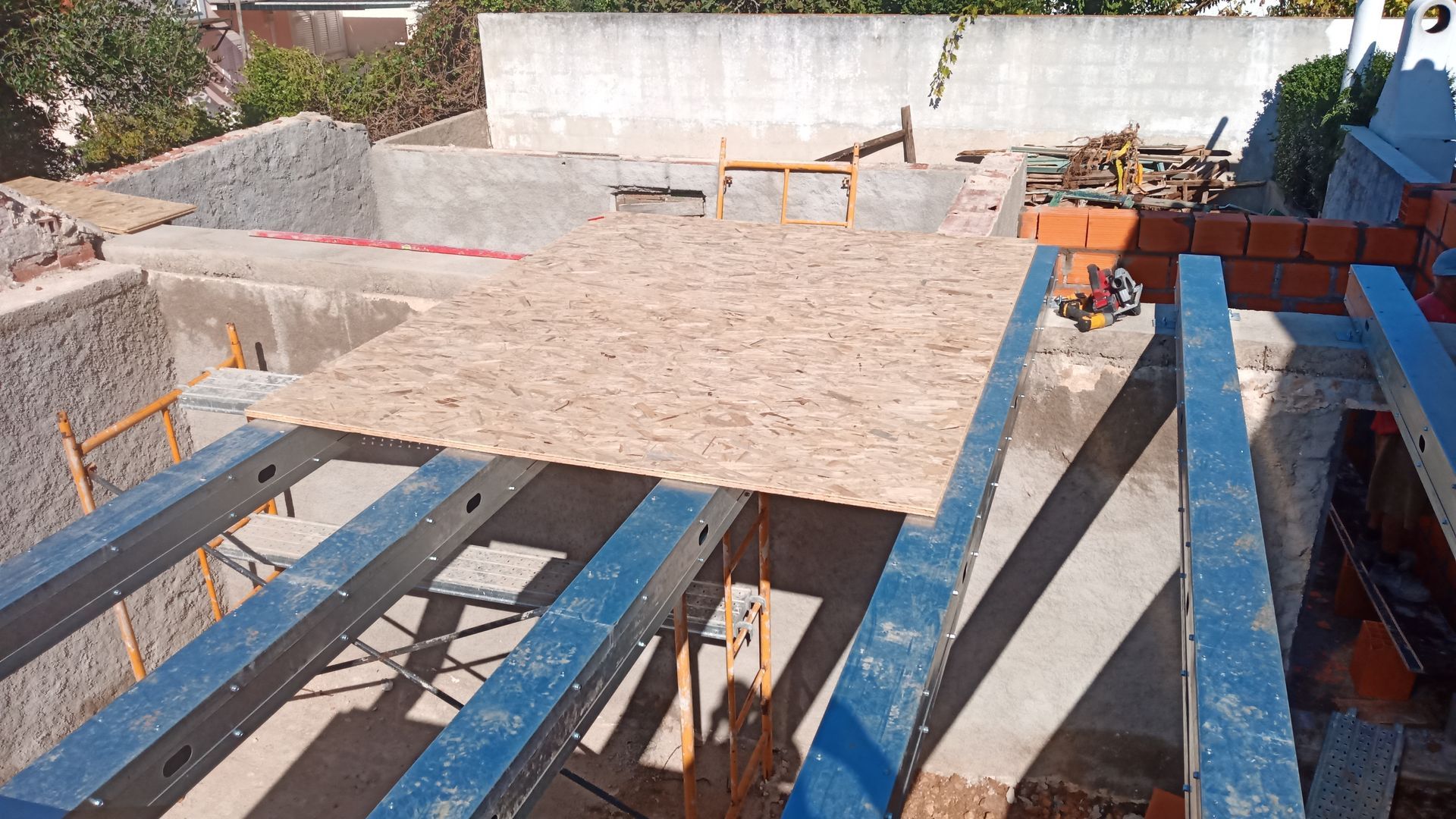 Canteiro de obras: painel OSB sobre vigas de aço azuis, com estruturas de tijolo e concreto.