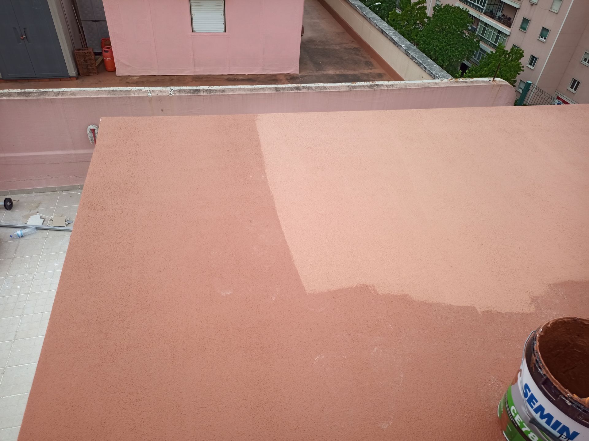 Telhado parcialmente pintado com tinta marrom-avermelhada; lata de tinta visível. Ambiente externo, luz do dia.