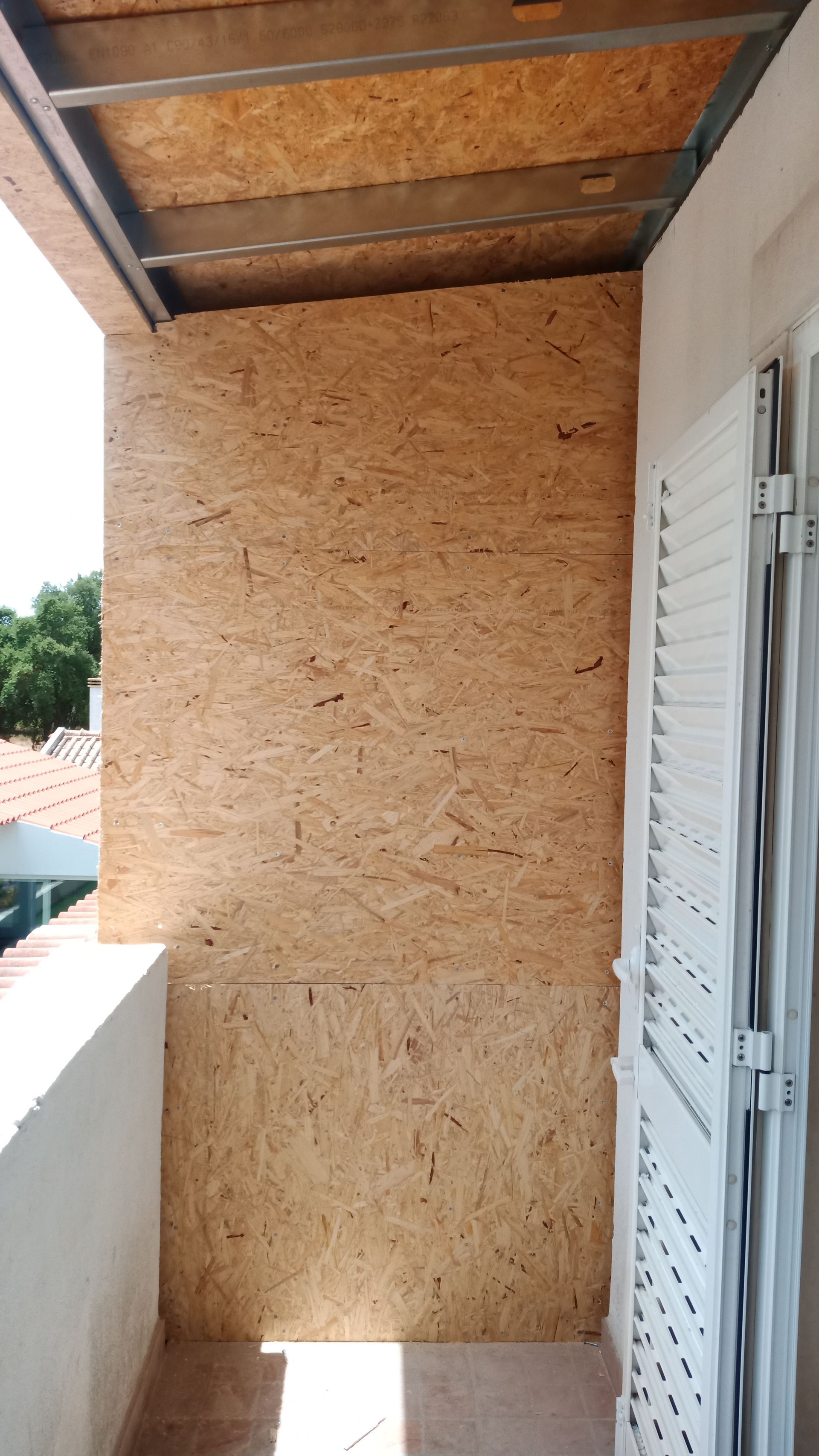 Painéis de OSB na parede da varanda, estrutura metálica no teto e porta branca com venezianas.
