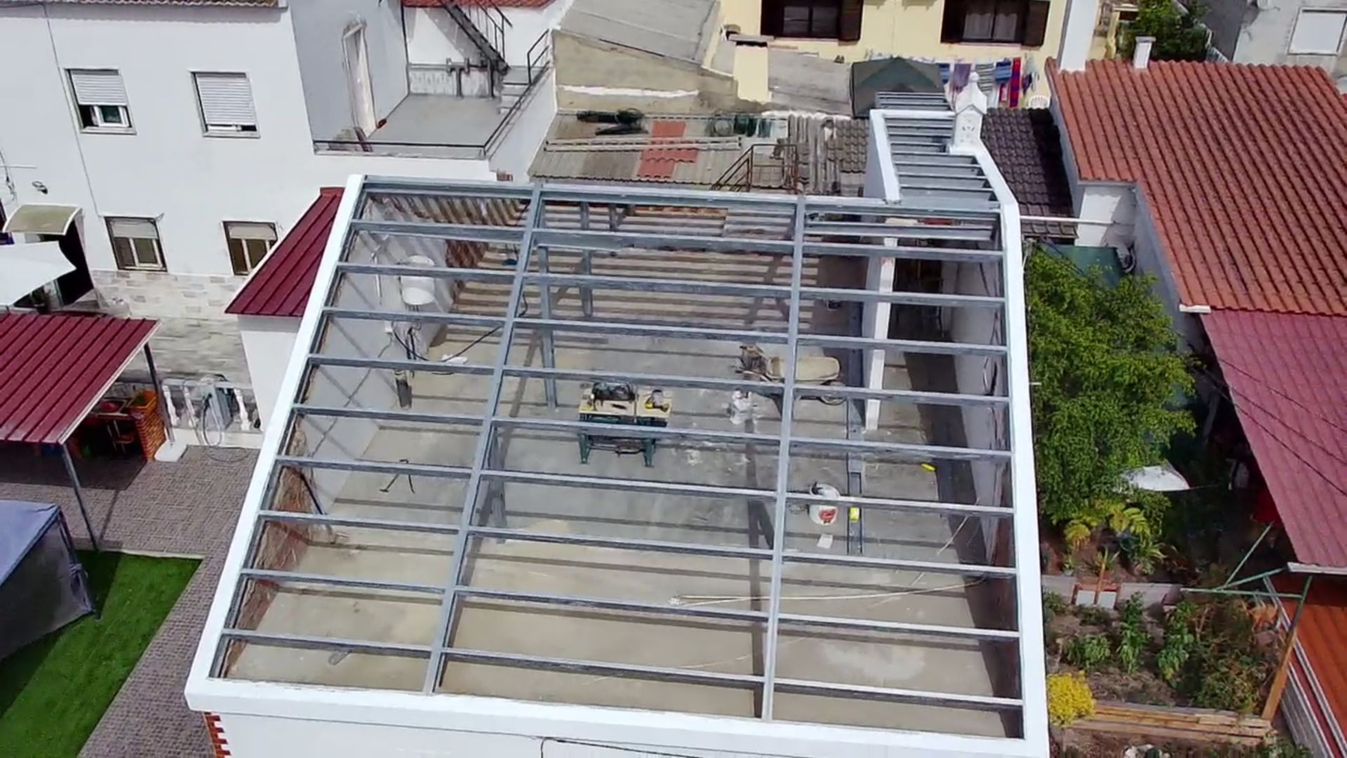 Vista aérea de uma estrutura metálica sobre o telhado plano de um edifício, cercada por outras construções.