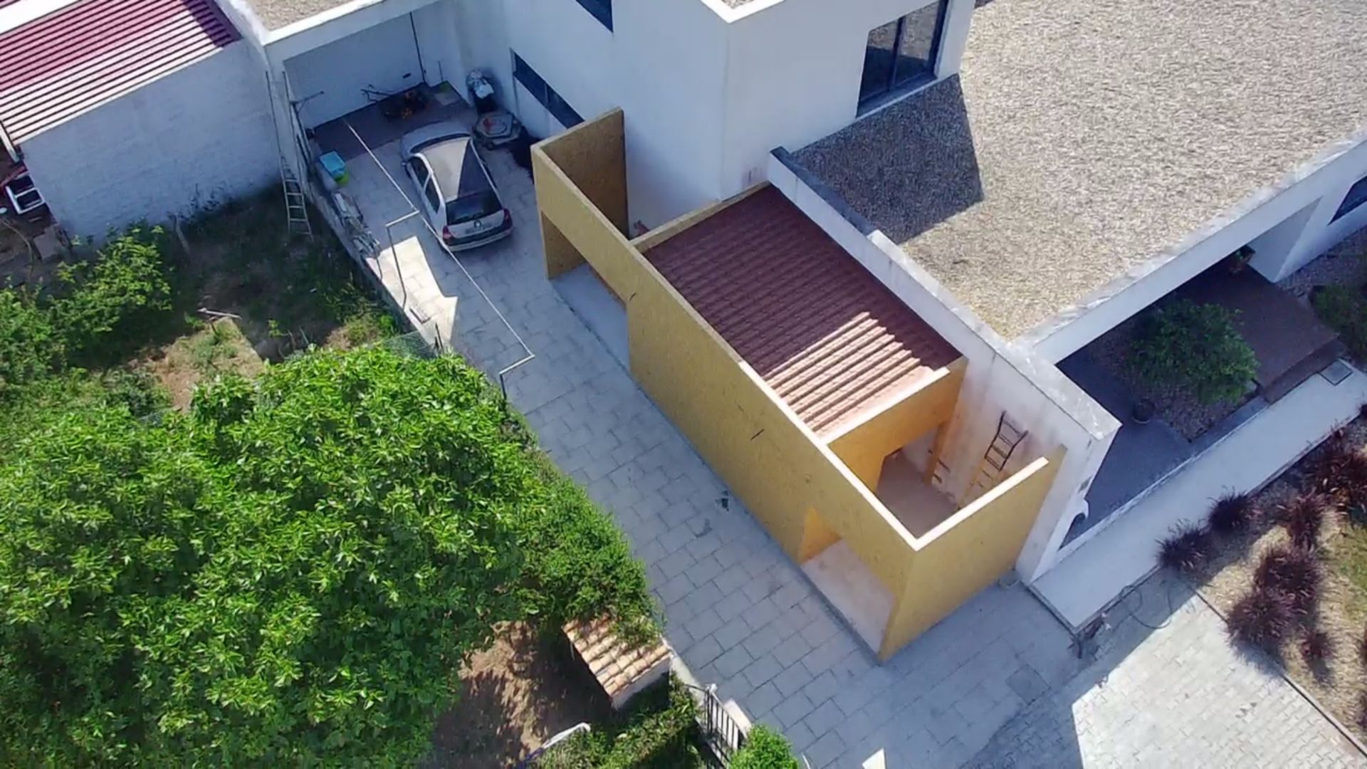Vista aérea de uma casa moderna com uma estrutura lateral amarela, entrada para carros e um carro estacionado na sombra.