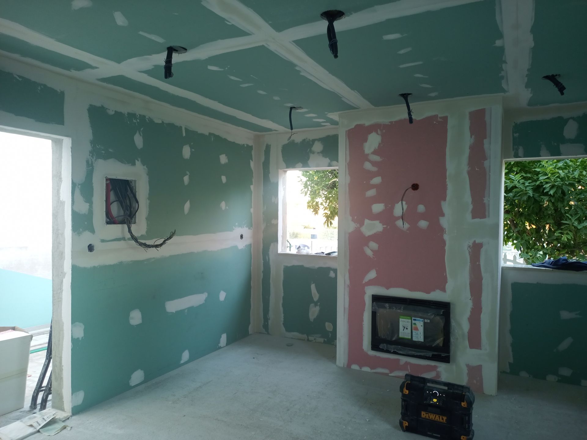 Quarto interno em construção, com paredes de gesso cartonado verdes e rosas, fiação exposta e lareira.