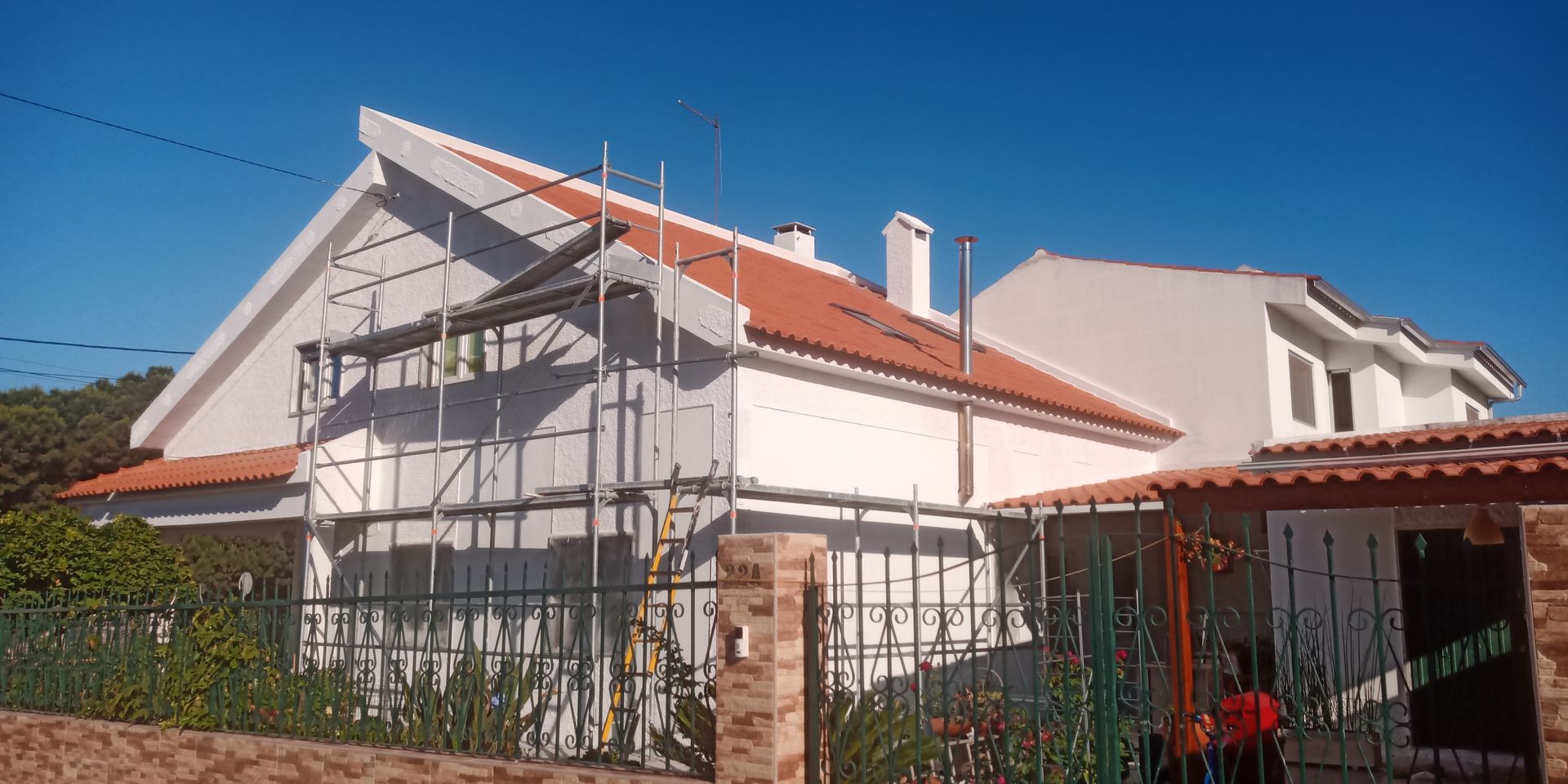 Casa branca em construção, com andaimes. Telhado de telha vermelha, cerca verde e céu azul claro.