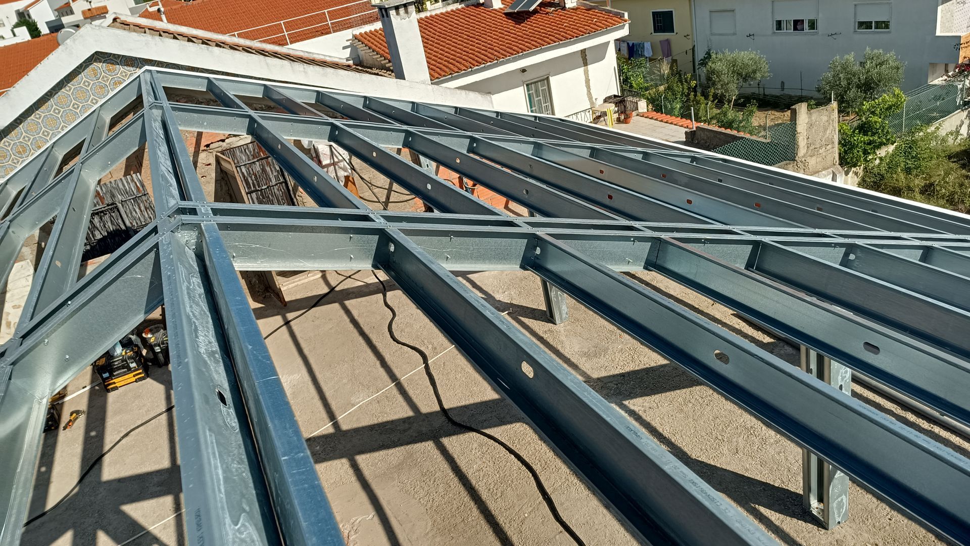 Estrutura metálica do telhado em construção, ao ar livre, com um telhado de telhas vermelhas ao fundo.
