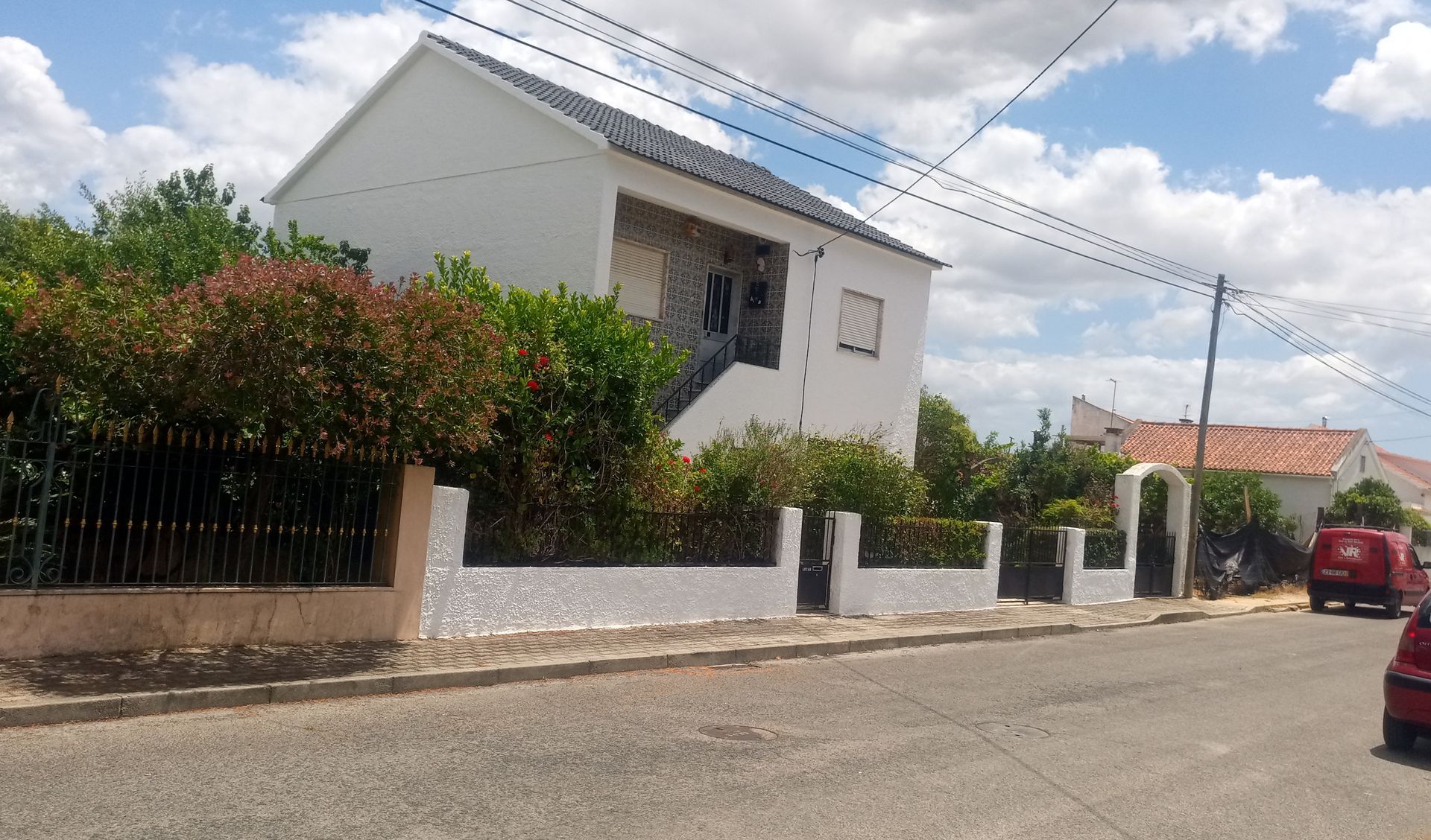 Casa branca de dois andares com arbustos, cerca preta e fios de energia em uma rua ensolarada.