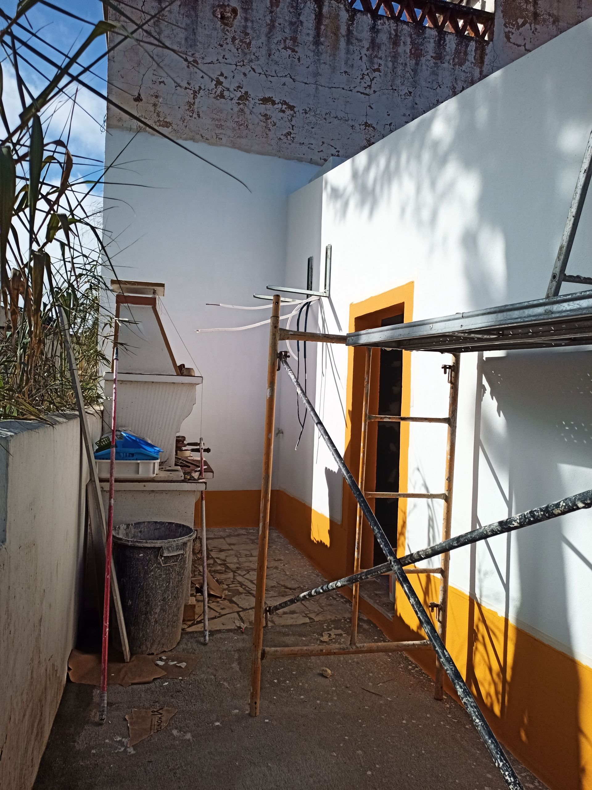 Exterior com andaimes, uma parede branca com detalhes em amarelo, uma porta e uma pequena estrutura de tijolos.