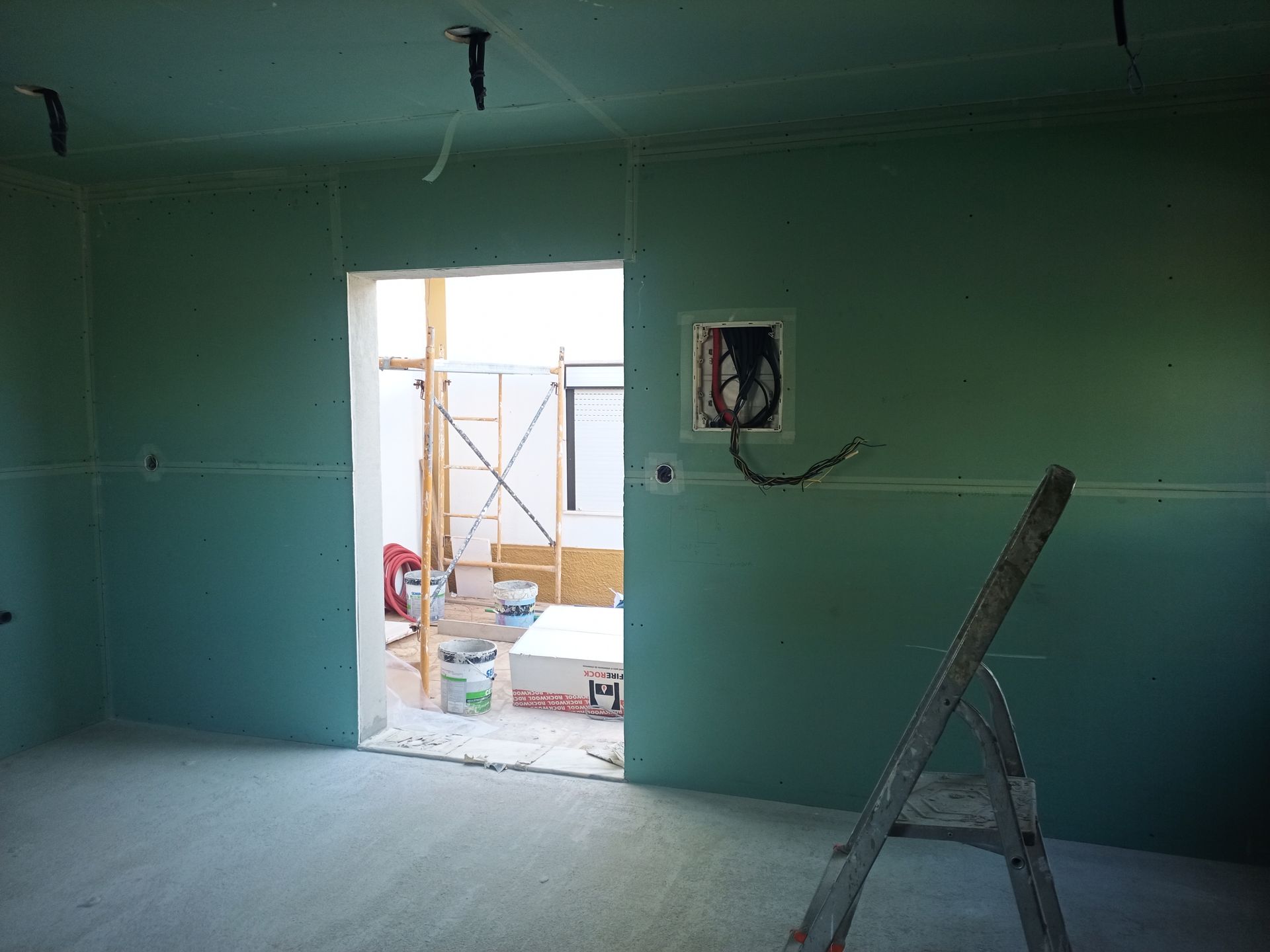Sala interna com parede de gesso verde, porta aberta e escada de mão. Fios e caixa de energia visíveis.