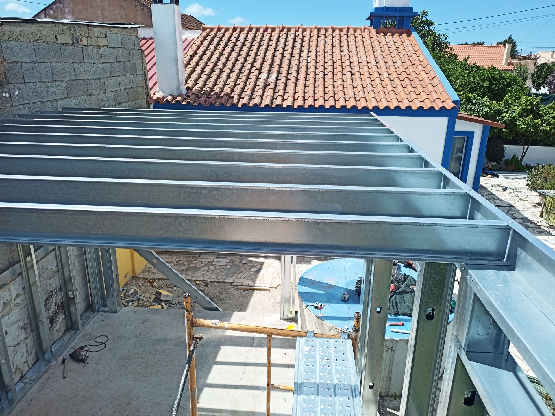 Estrutura metálica para cobertura, com materiais de construção e andaimes visíveis, sob um céu claro.