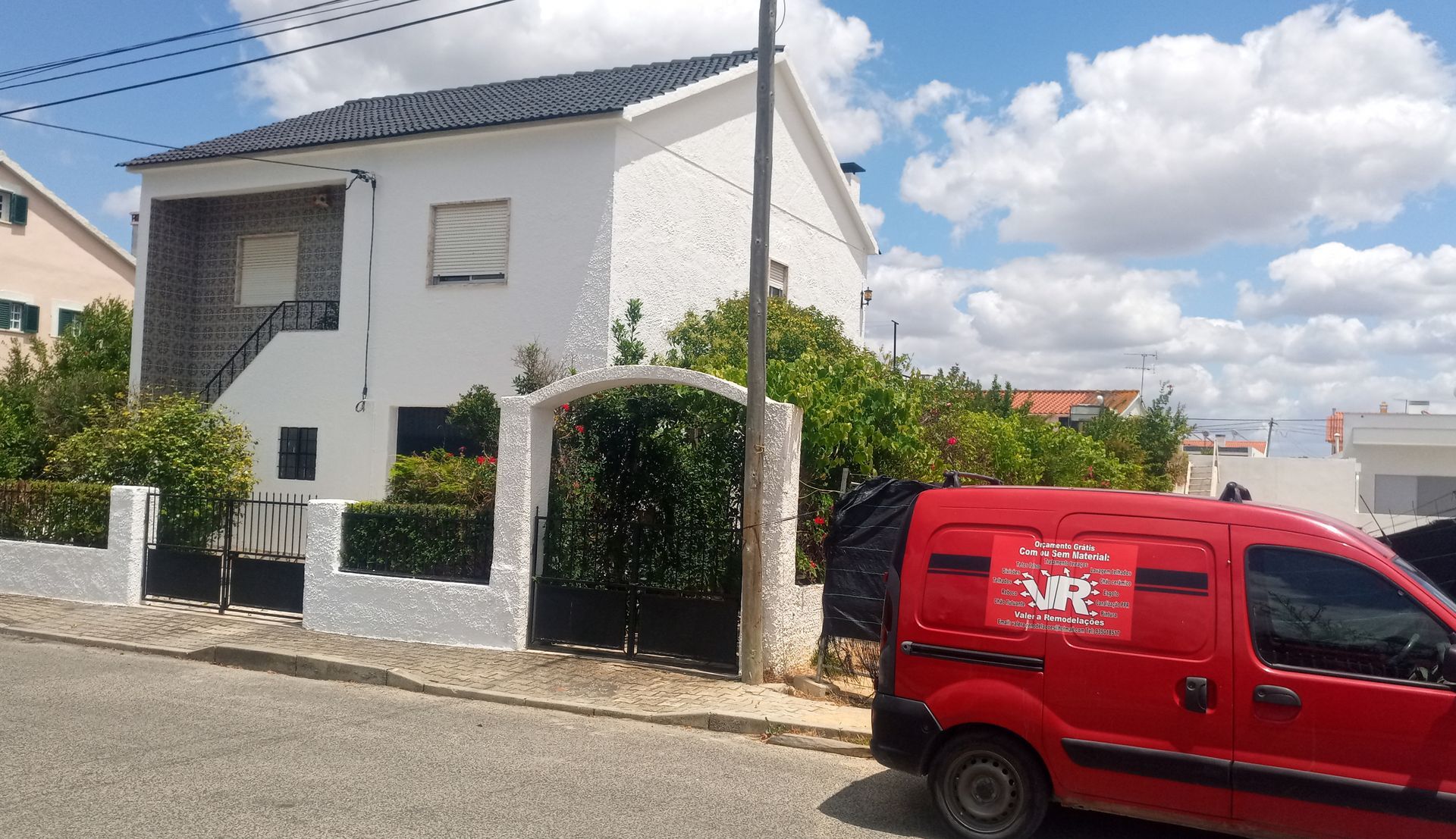Casa branca com portão preto, ao lado de uma van vermelha. Dia ensolarado, céu azul.