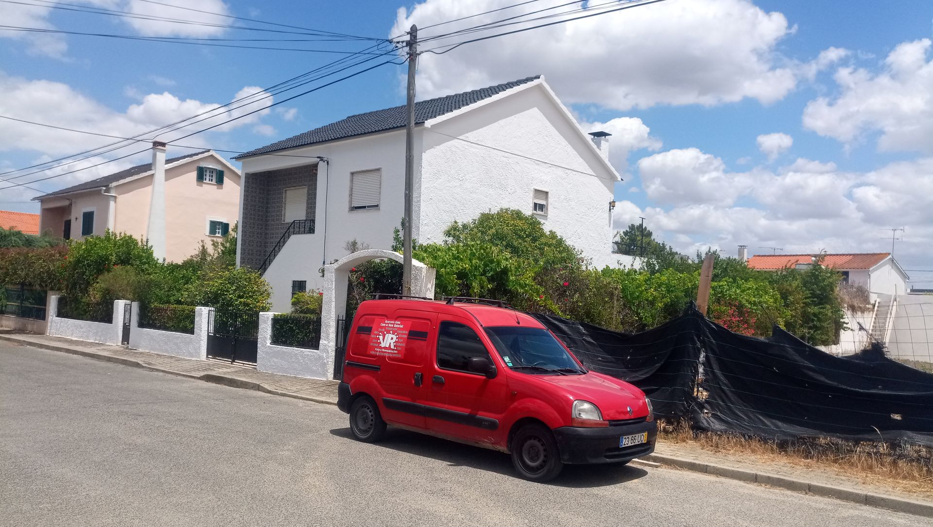 Uma van vermelha estacionada na rua em frente a uma casa branca com vegetação. Céu azul acima.