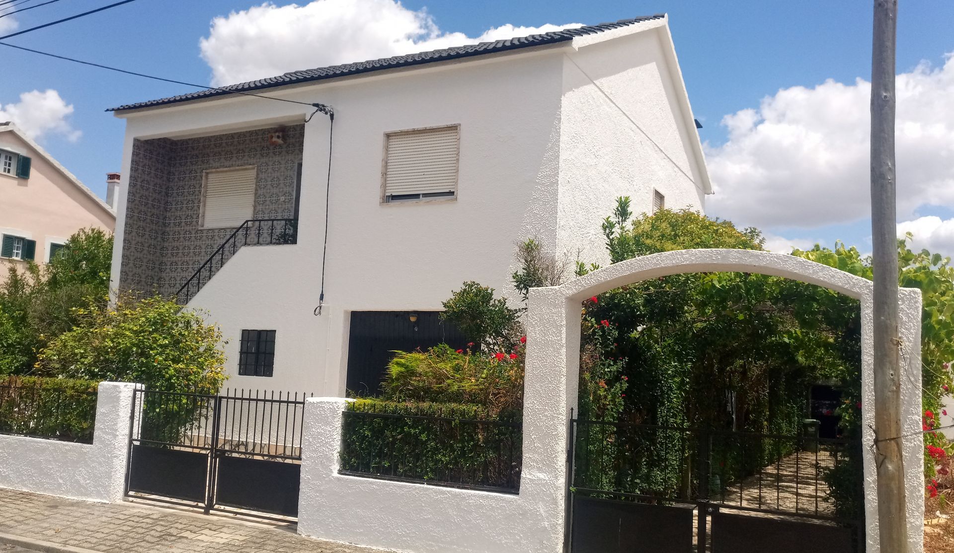 Casa branca de dois andares com um pequeno jardim, atrás de uma cerca e portão brancos, sob um céu azul.