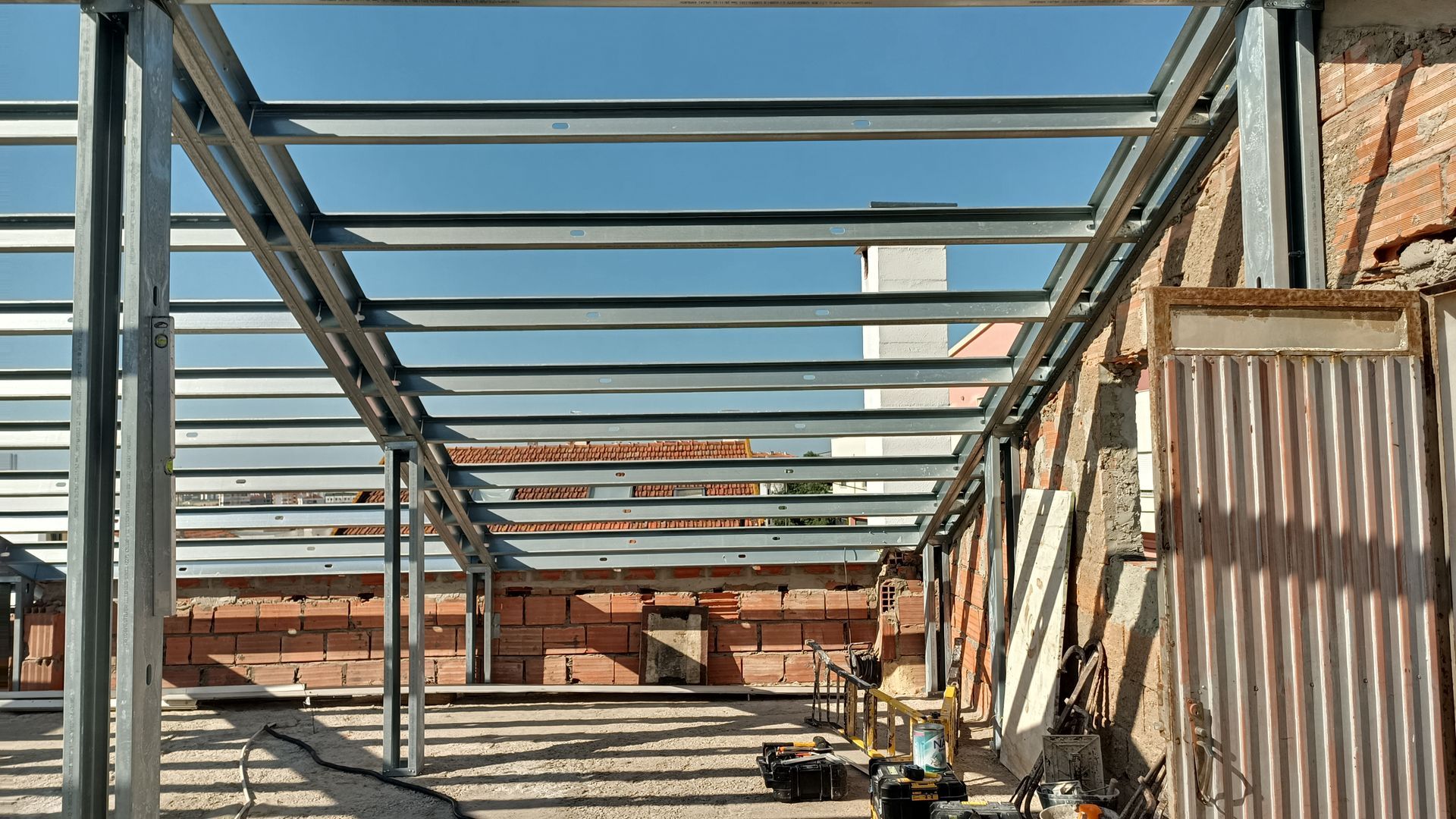 Telhado com estrutura de aço em construção, visível contra as paredes de tijolo e o céu azul.