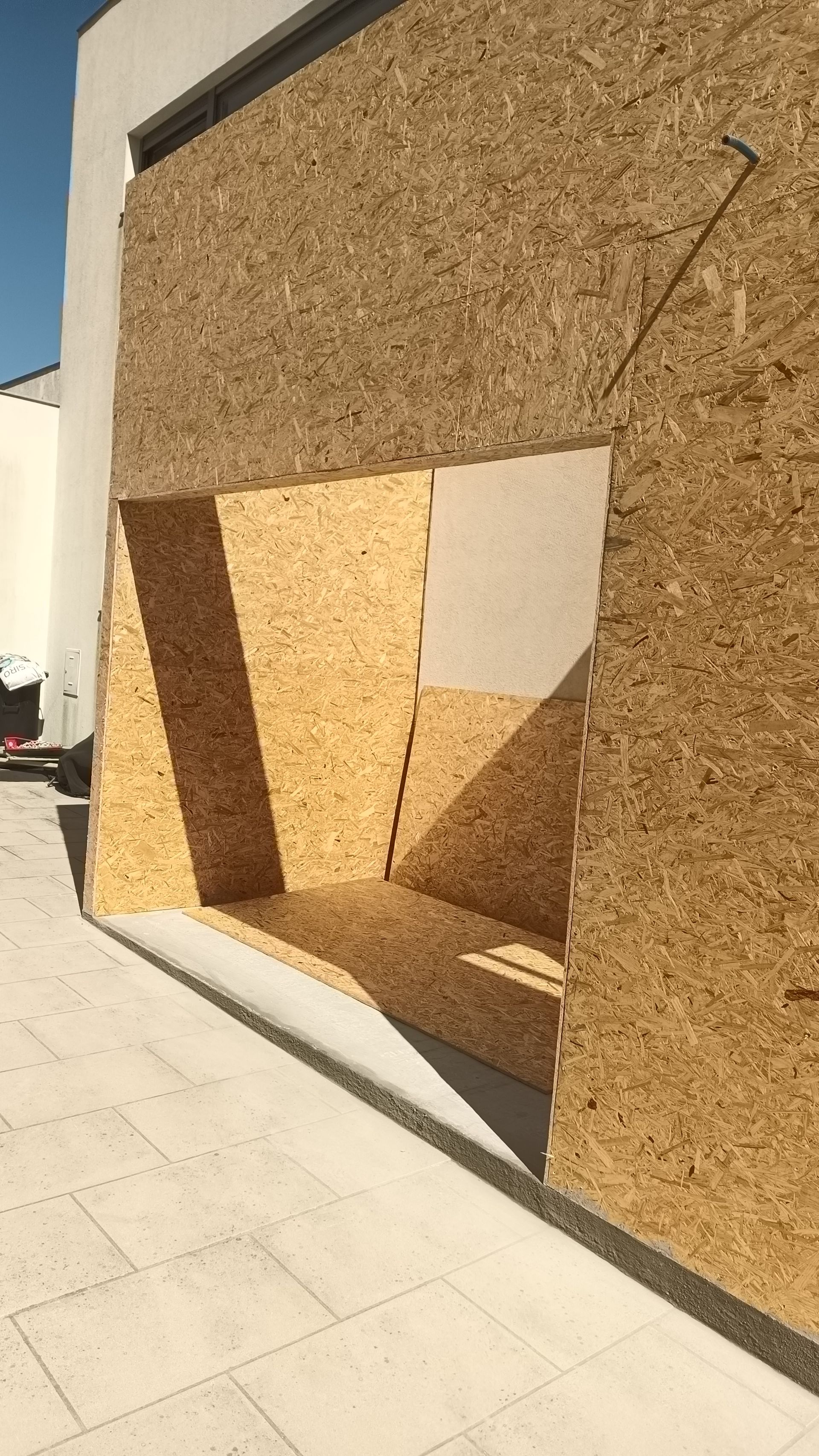 Parede externa com placas de OSB cobrindo a abertura de uma janela, projetando sombras na superfície.